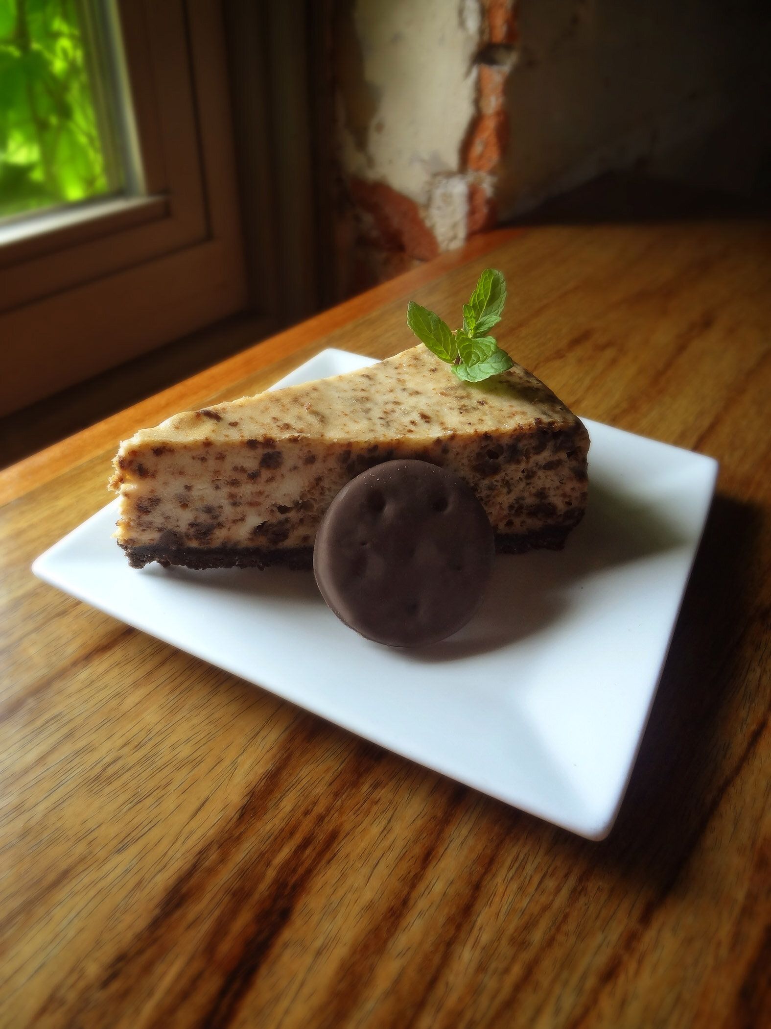 A slice of chocolate chip cheesecake topped with mint on a square white plate, with a cookie next to it on wood grain.