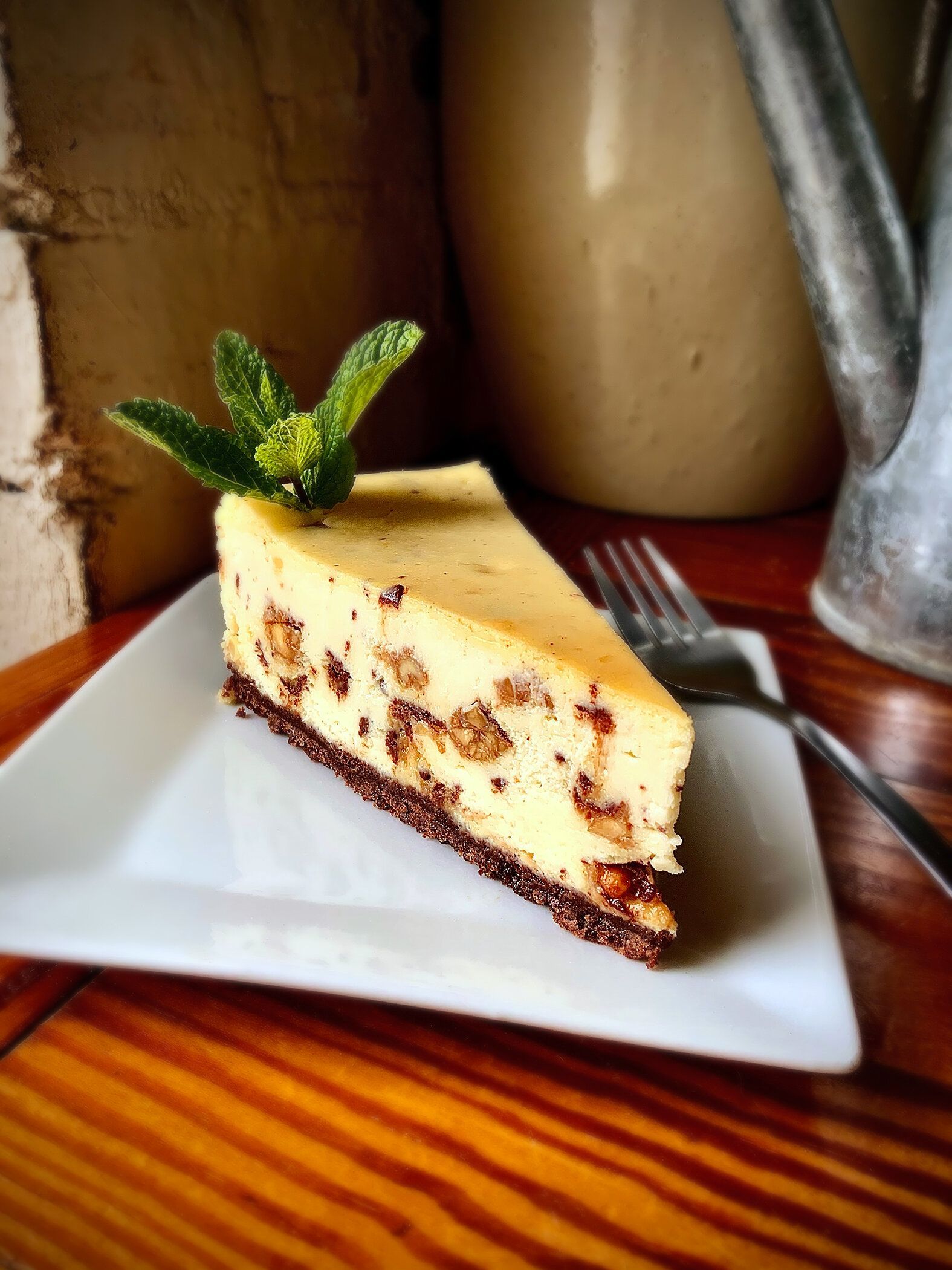 A slice of cheesecake with a chocolate crust and inclusions, garnished with fresh mint on a white plate by a metal pitcher.