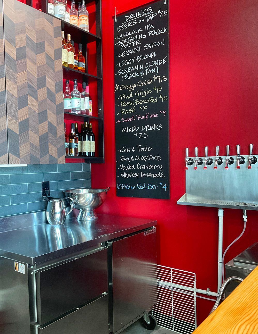 A bar area featuring a chalkboard menu on a bright red wall, a stainless steel counter, shelves of liquor, and beer taps.
