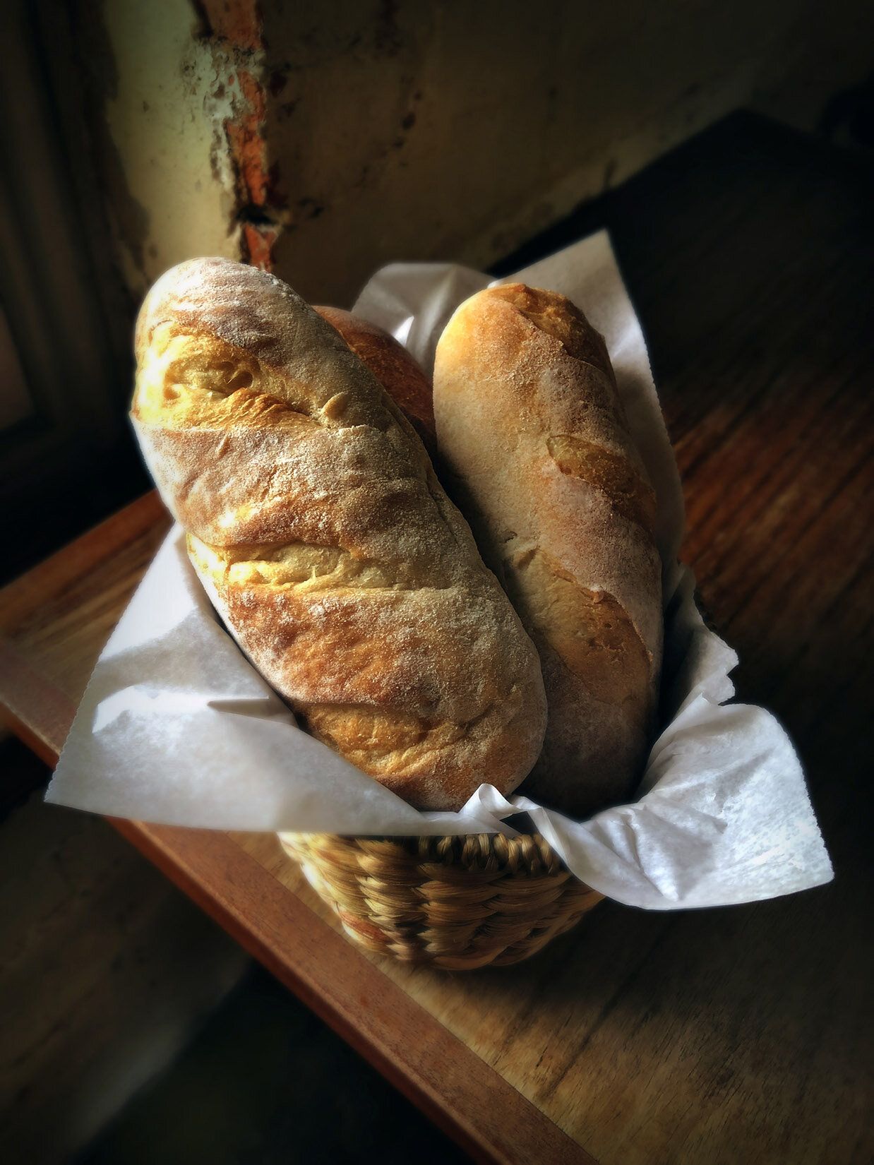 A wicker basket lined with white paper holds two crusty loaves of bread on a wooden surface.