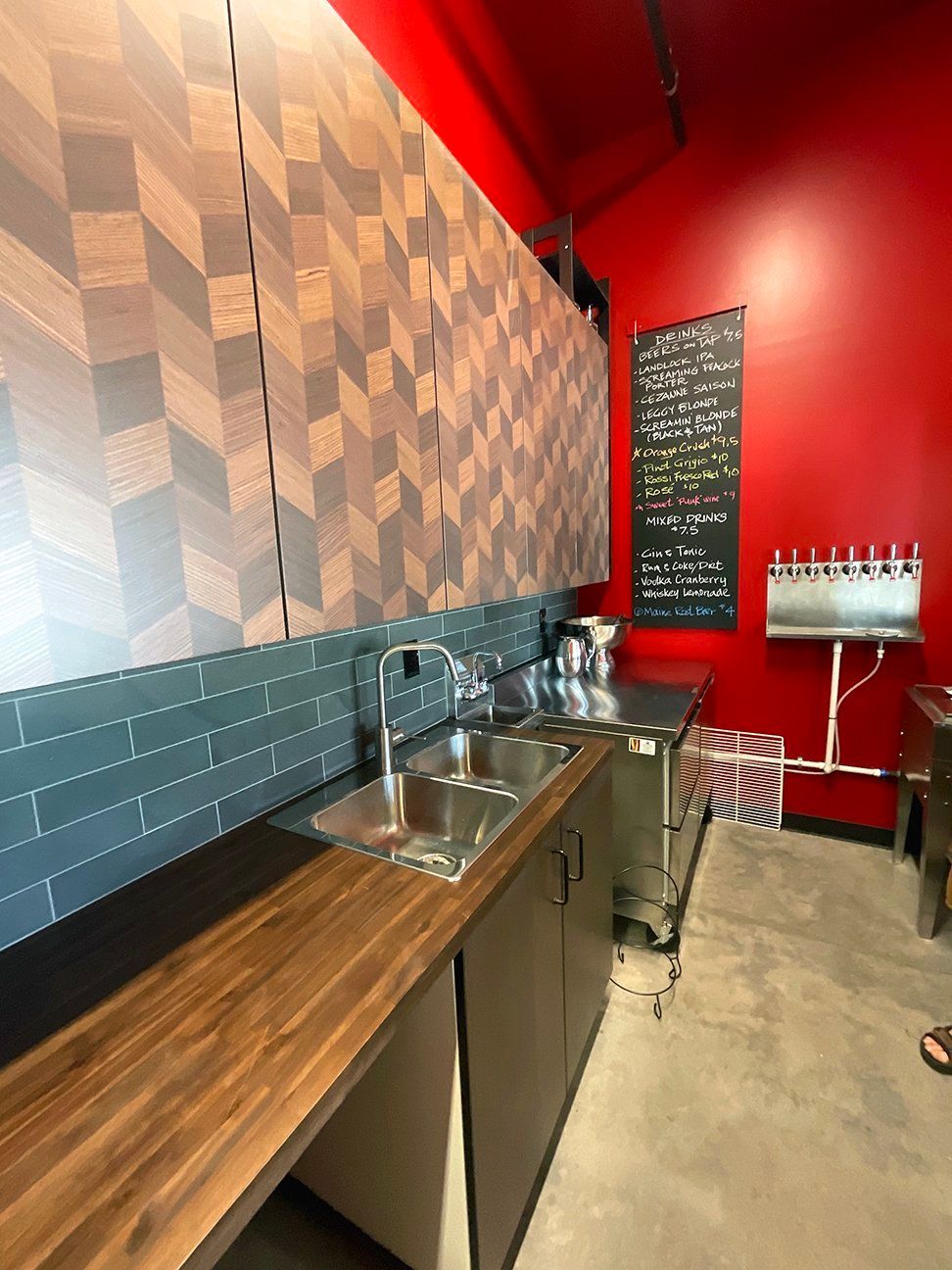 A kitchen area with a wooden countertop, a double sink, teal subway tile backsplash, chevron cabinets, and a red wall.