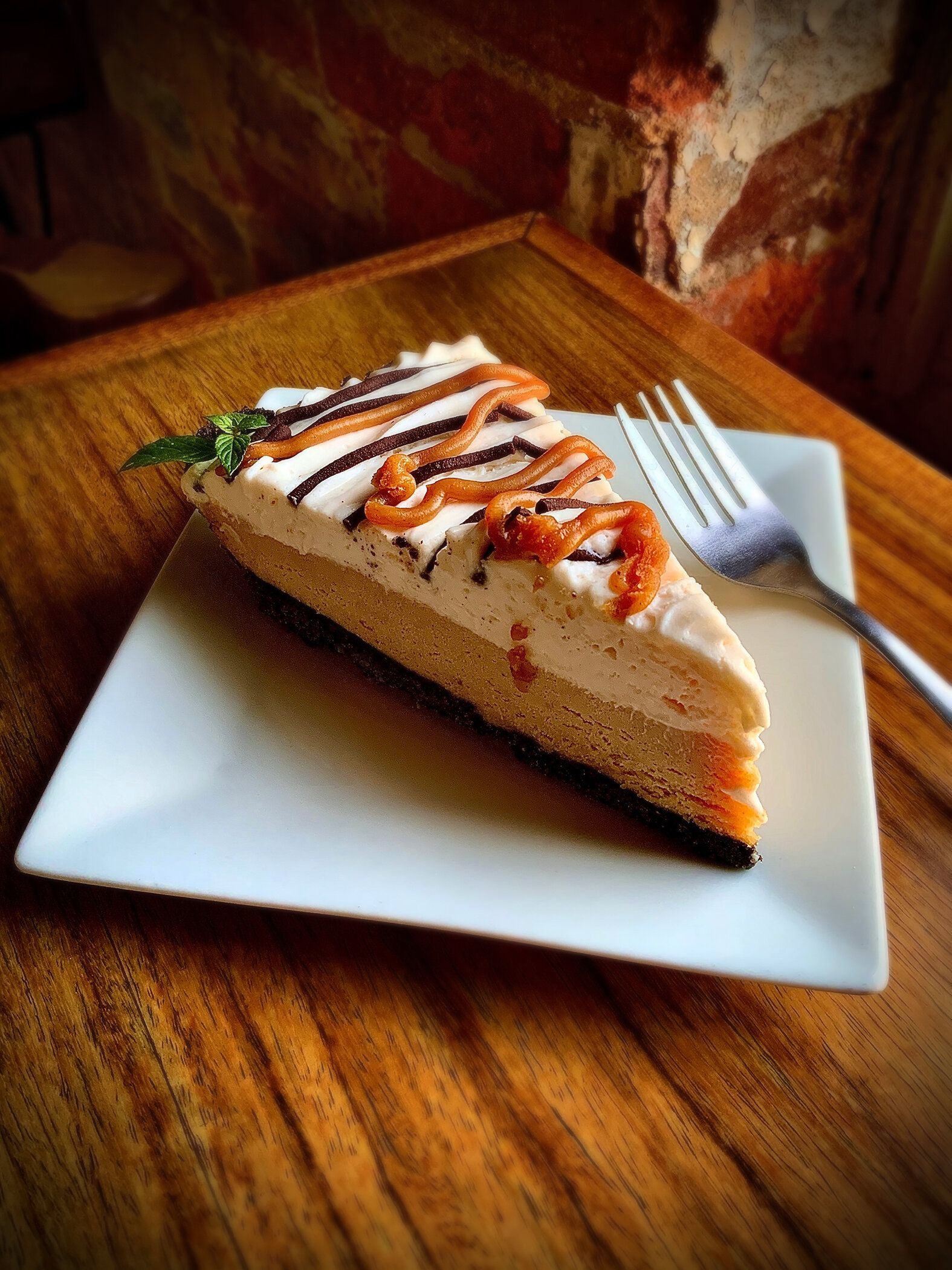 A slice of cheesecake with caramel and chocolate drizzle on a white plate next to a fork on a wooden table.