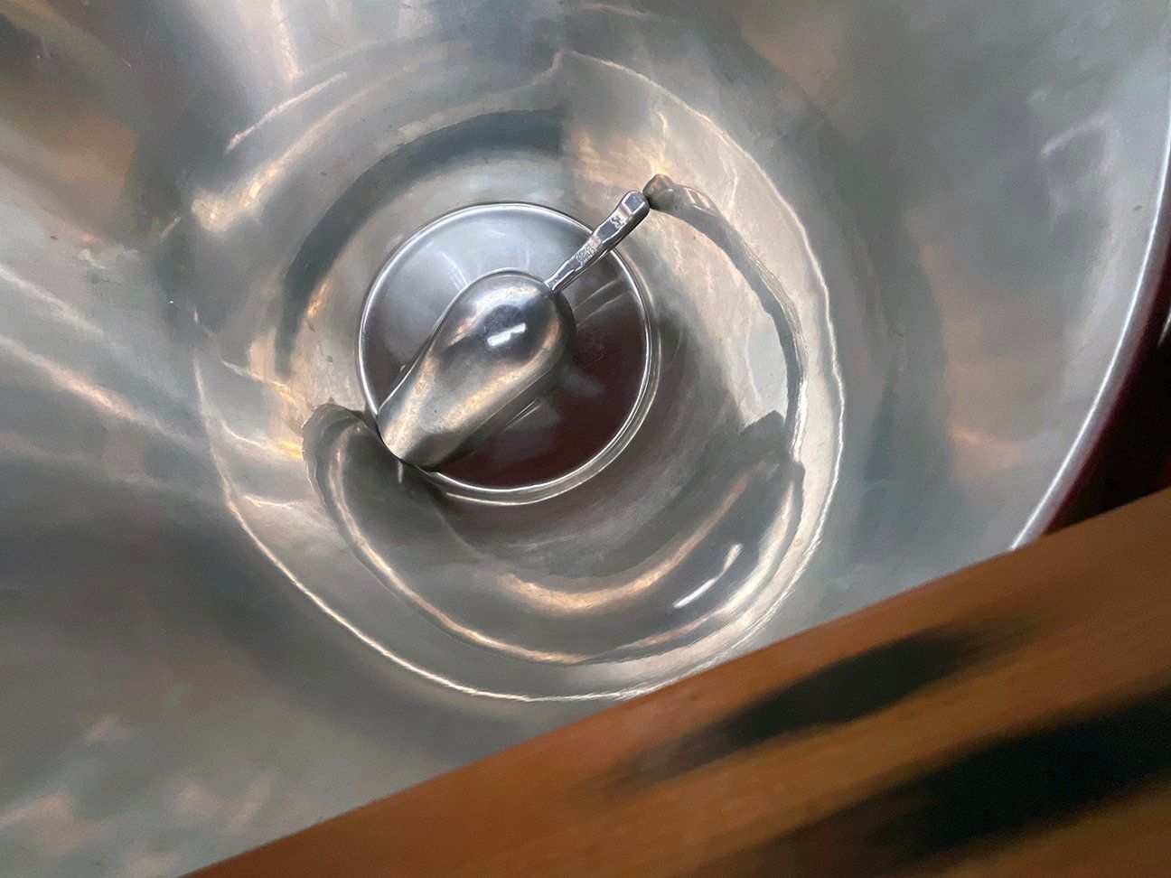 A metal scoop resting inside the polished, funnel-shaped stainless steel hopper of a food processing machine.