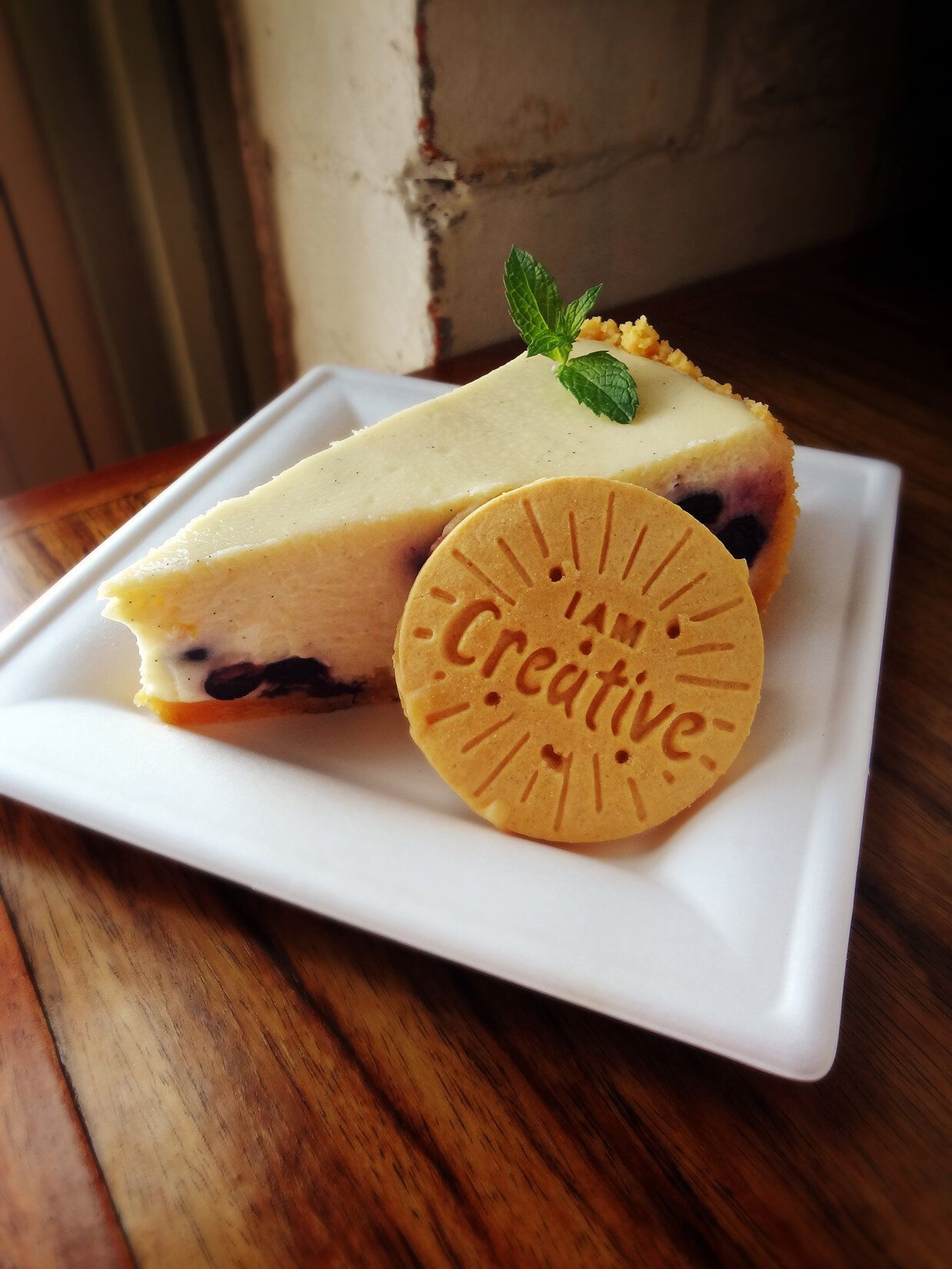 A slice of blueberry cheesecake garnished with a mint sprig and a round cookie labeled 