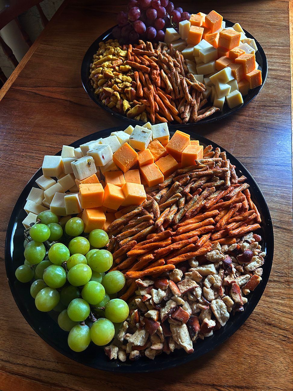 Two round serving platters on a wooden table, filled with cubed cheese, green and red grapes, pretzels, and chopped nuts.