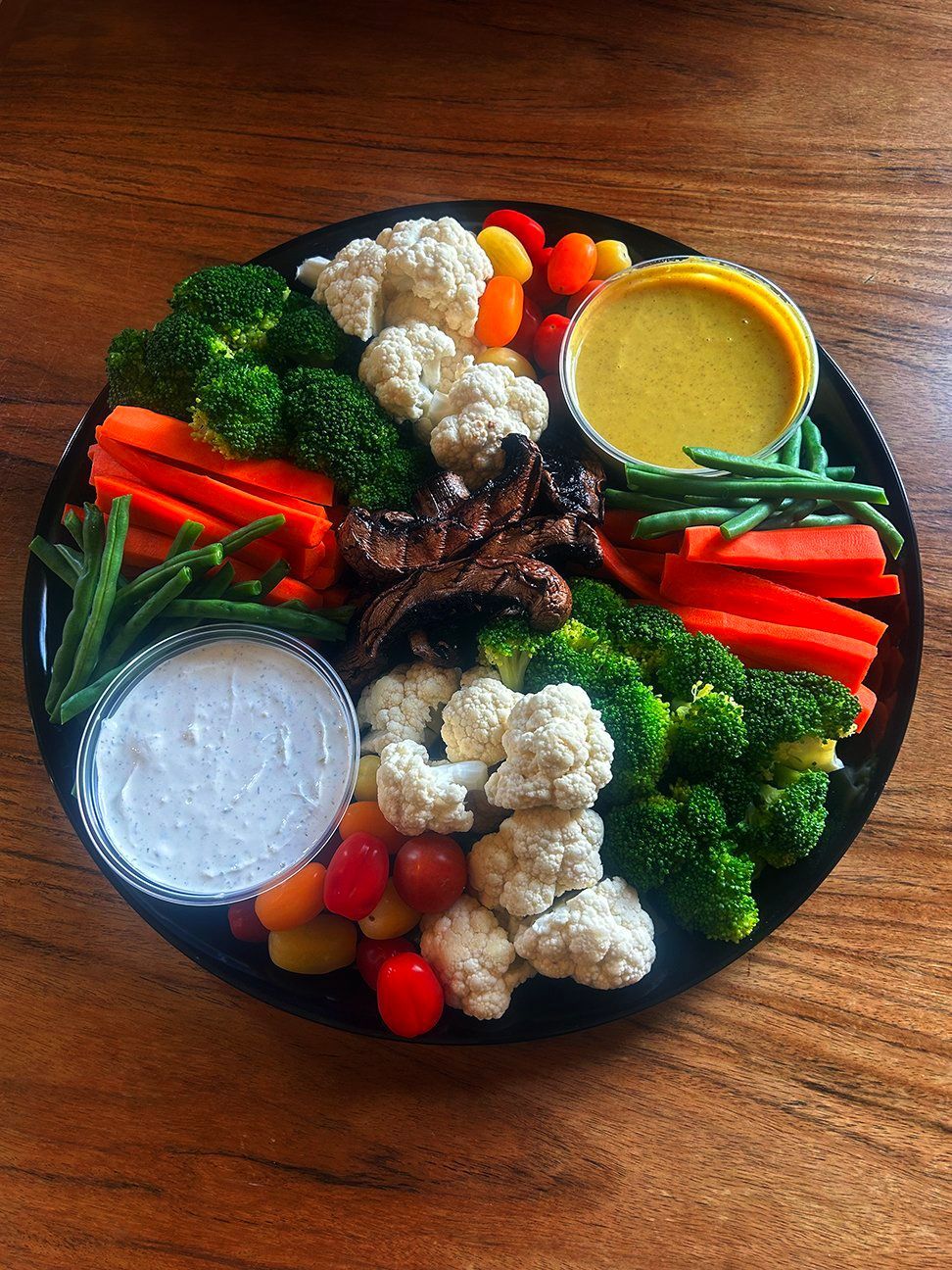 A platter of fresh vegetables including broccoli, cauliflower, carrots, green beans, tomatoes, and mushrooms with two dips.