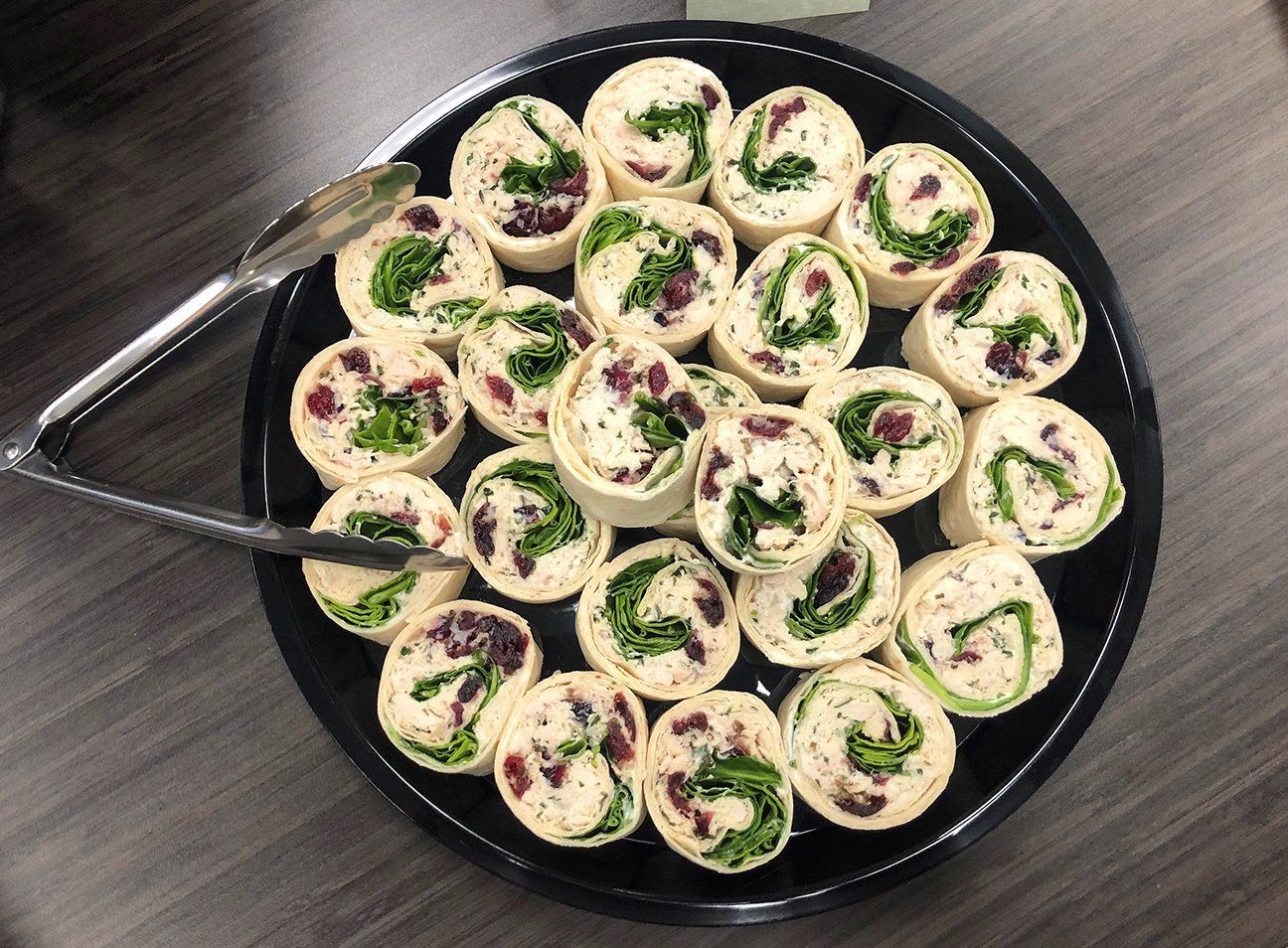 A black tray holds a circular arrangement of sliced tortilla pinwheels with greens and dried fruit, served with tongs.