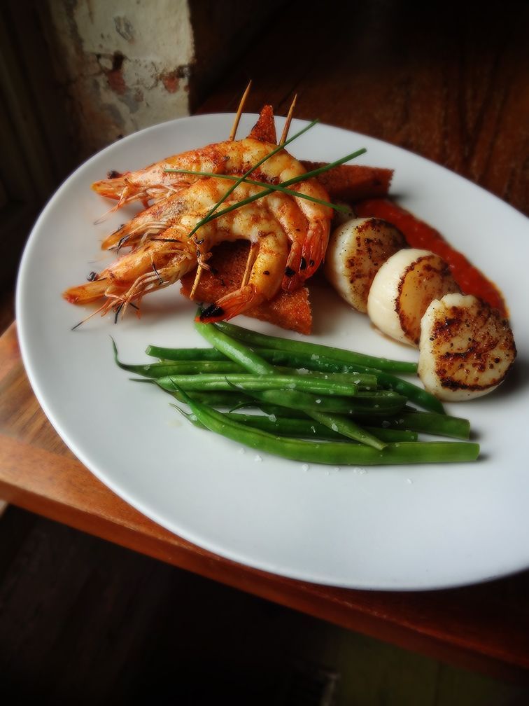 A white plate holds seared scallops, grilled shrimp on skewers, and green beans, all served with a side of red sauce.