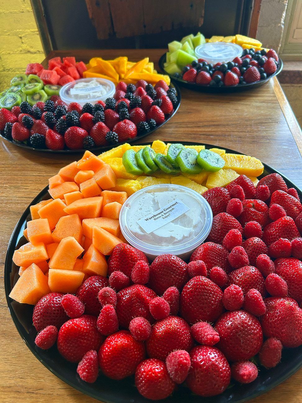 Three round platters of assorted fresh fruit, including strawberries, cantaloupe, pineapple, kiwi, and dip, on a table.