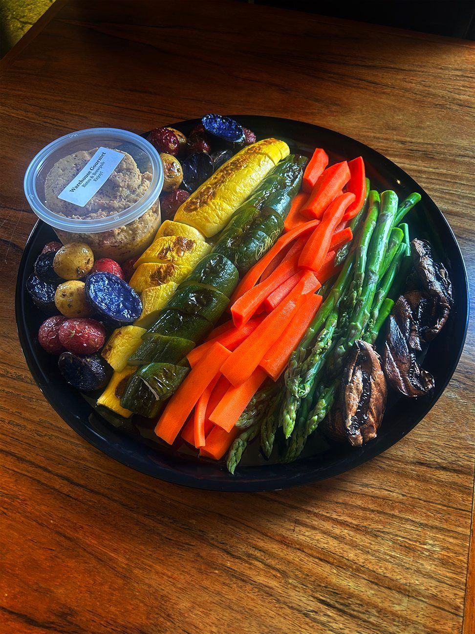 A round platter on a wooden table holds grilled zucchini, carrots, asparagus, roasted mushrooms, potatoes, and a dip.
