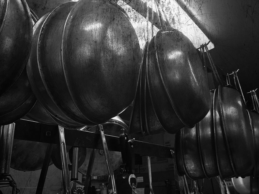 A low-angle, black-and-white view of several large, round metal pots hanging from a rack in a kitchen.