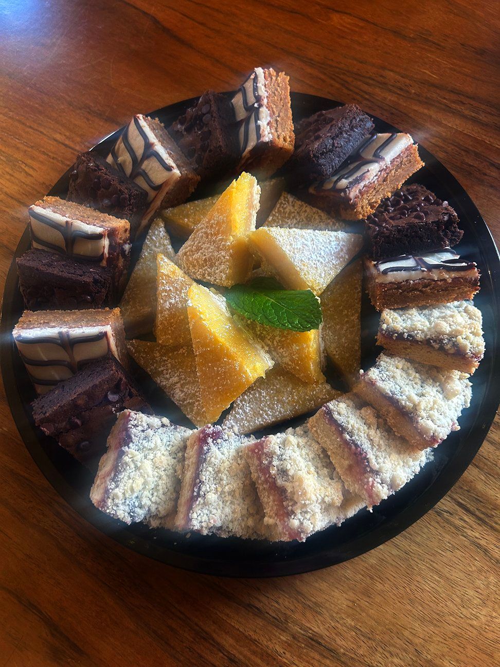 A circular platter on a wooden table holds assorted bar cookies, including chocolate glazed, yellow, and crumb-topped.