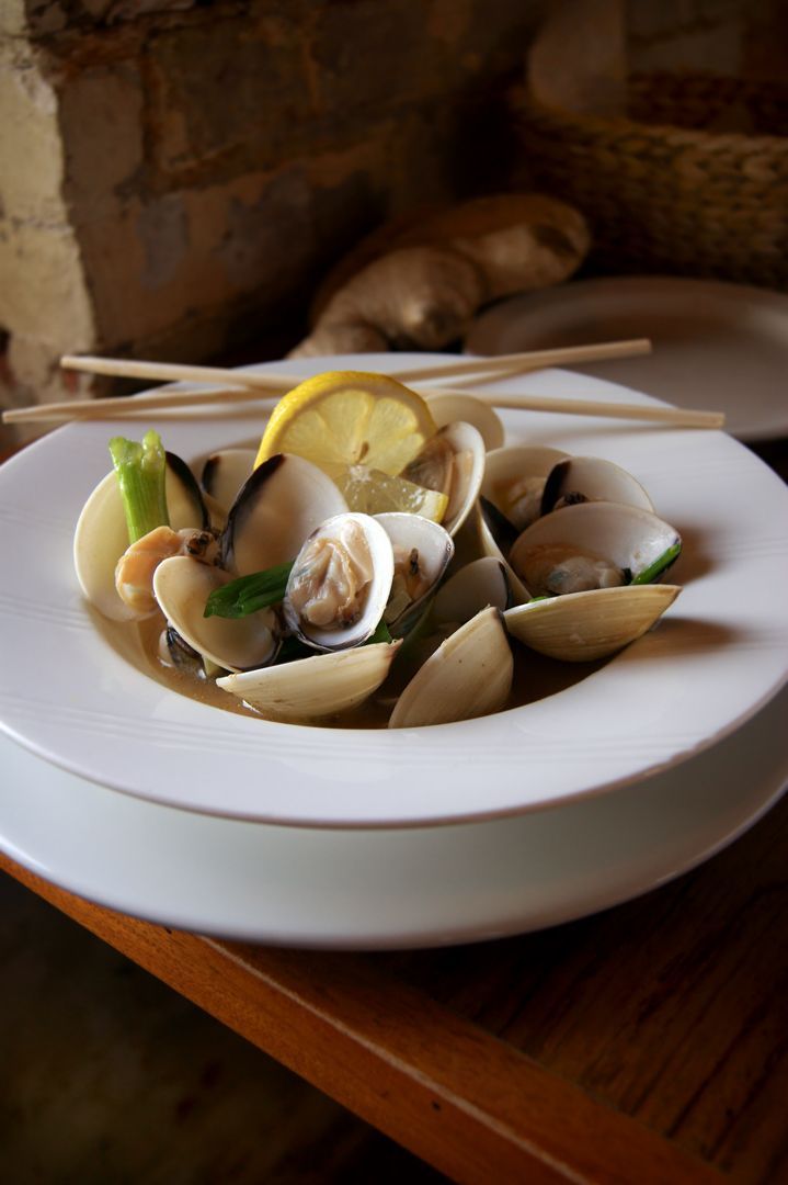 A bowl of steamed clams in a light broth, garnished with a lemon slice and herbs, served with chopsticks on a wooden table.