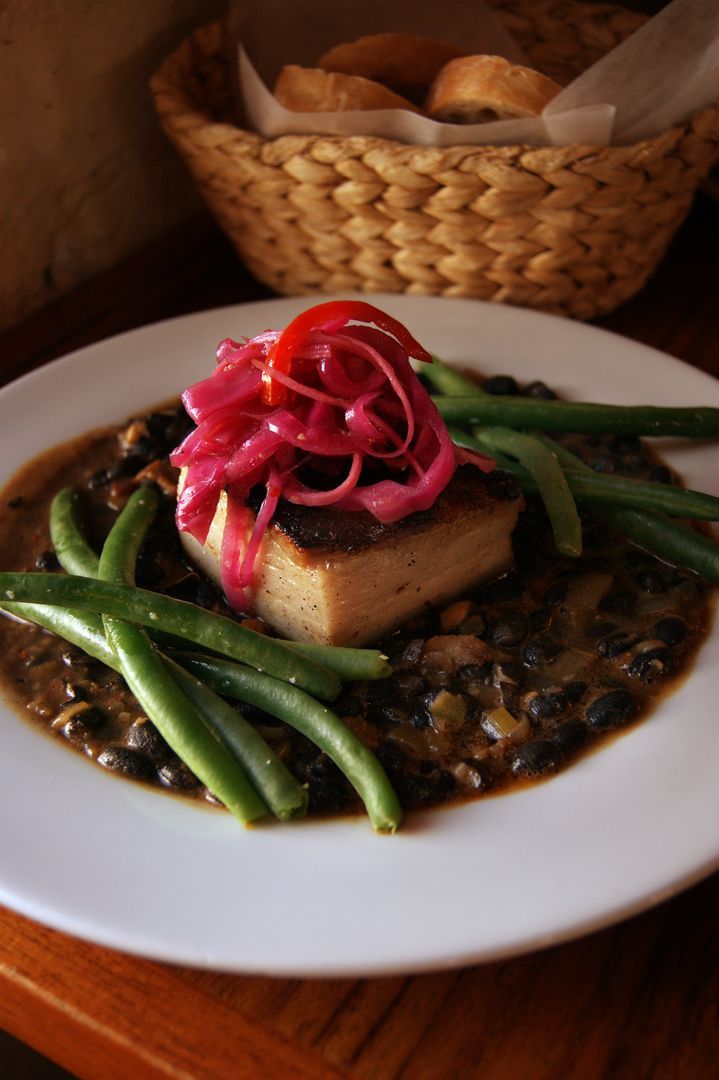 A plated meal of seared fish on black beans with green beans and pickled red onions, with a bread basket in the background.