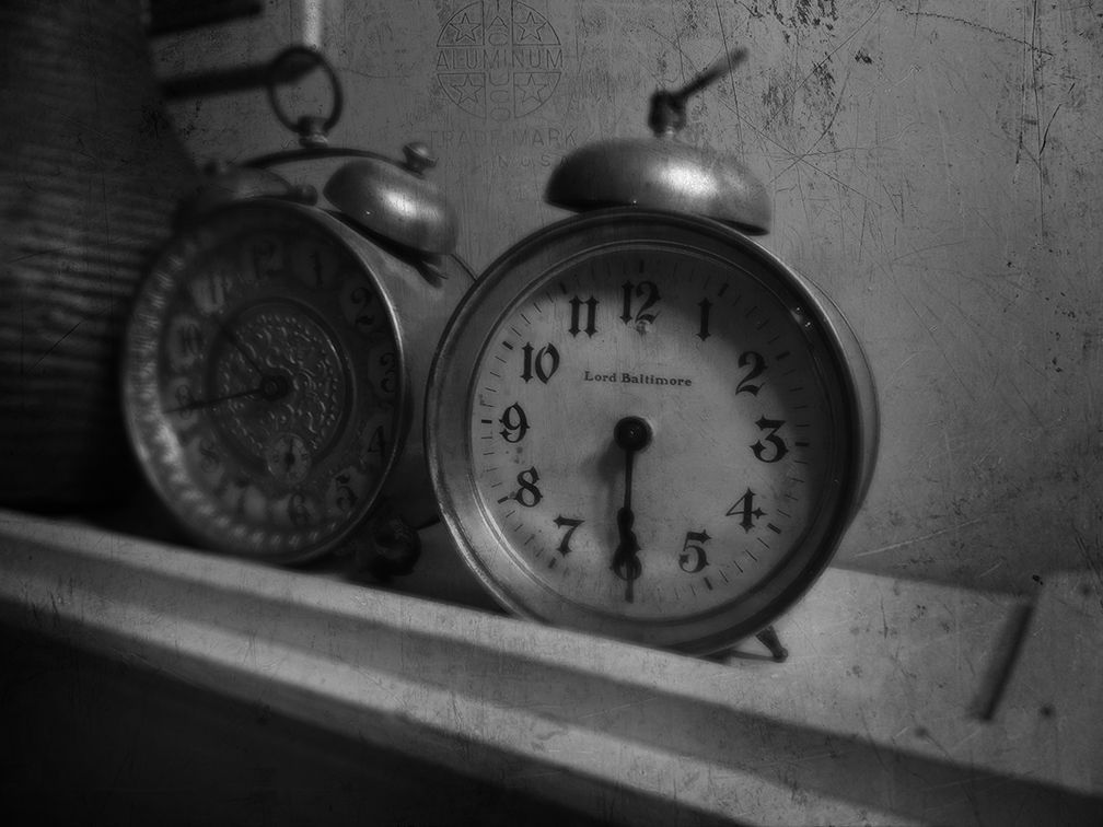 Two vintage analog alarm clocks sit side-by-side on a white shelf in a grayscale, dimly lit room.