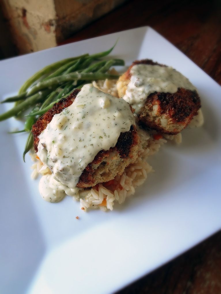 Two breaded crab cakes topped with creamy sauce served over rice with a side of green beans on a white square plate.