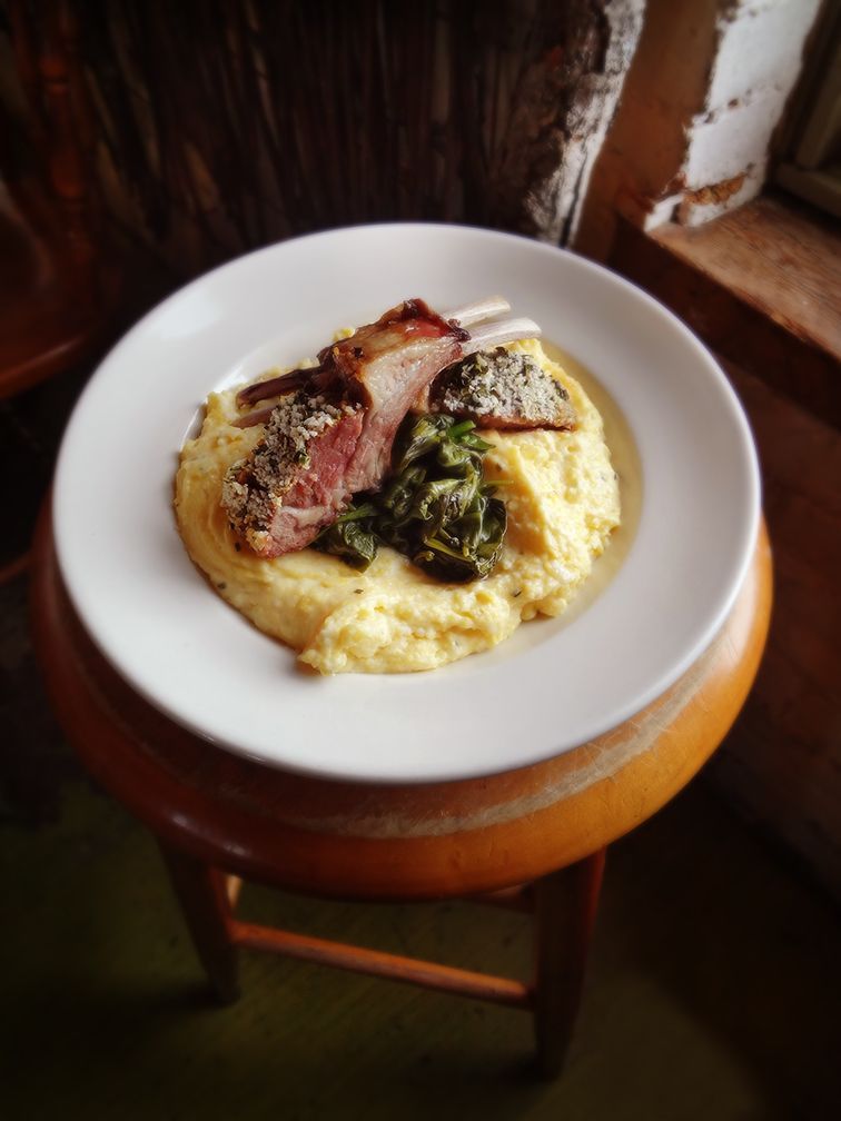 A plate featuring a lamb chop, cooked greens, and creamy polenta, resting on a wooden stool.