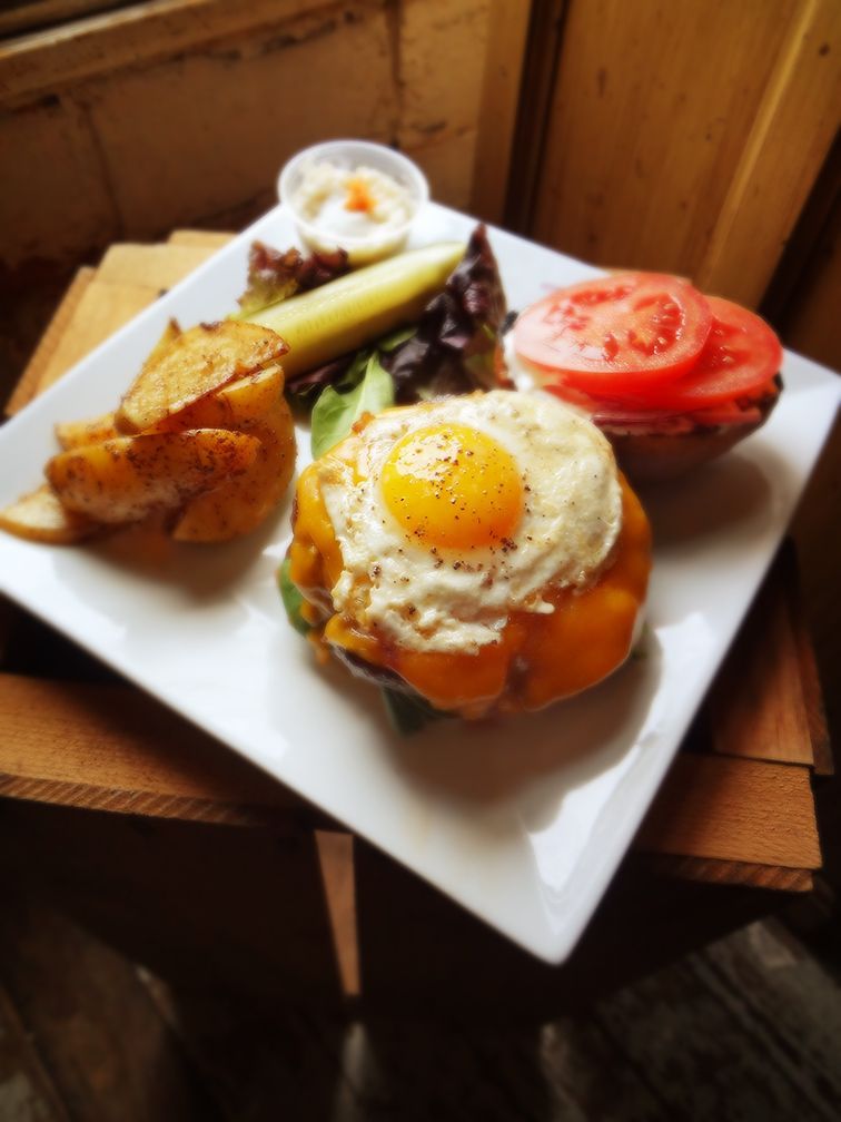 A burger topped with a sunny-side-up egg and melted cheese, served with potato wedges, a pickle, and tomato slices.