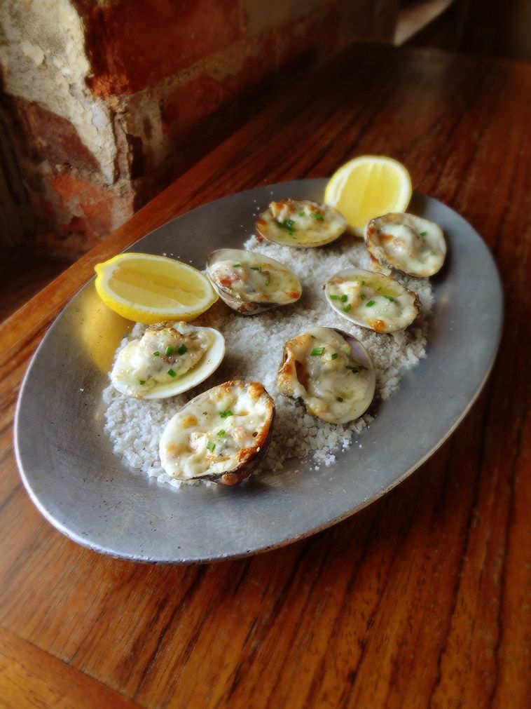Baked clams with herbs on a bed of rock salt on an oval metal platter with two lemon wedges, set on a wooden table.