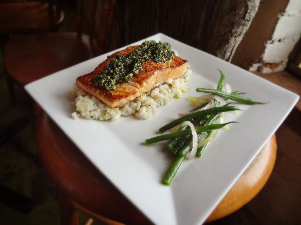 A pan-seared salmon fillet topped with pesto, served on a bed of herb risotto with a side of sautéed green beans and onions.