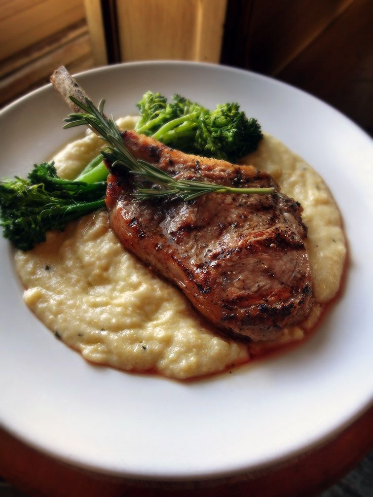 A grilled lamb chop with a rosemary sprig, served on a bed of creamy polenta with steamed broccoli on a white plate.