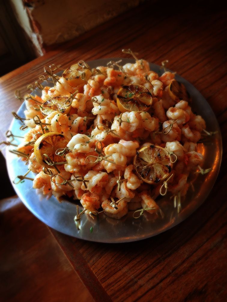 A metal platter holds many shrimp skewers interspersed with grilled lemon slices, set on a wooden table.