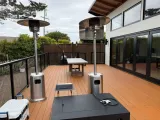 Two outdoor patio heaters on a wooden deck with a table, next to a house with large windows.
