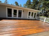 Wooden deck in front of a light gray house with white doors and railing, surrounded by trees.