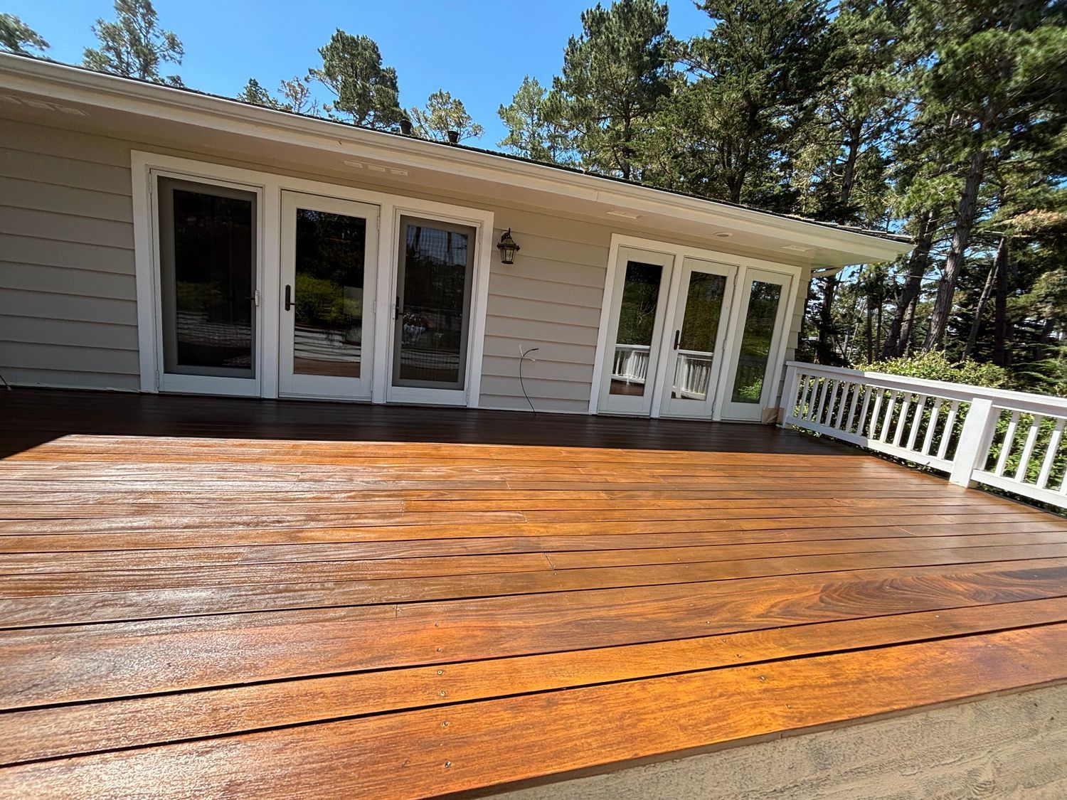 Wooden deck in front of a light gray house with white doors and railing, surrounded by trees.