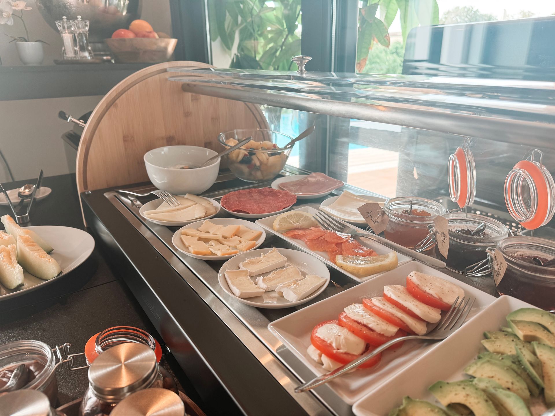 Breakfast buffet with various foods on display, including cheese, tomatoes, salmon, and avocado, in a bright setting.
