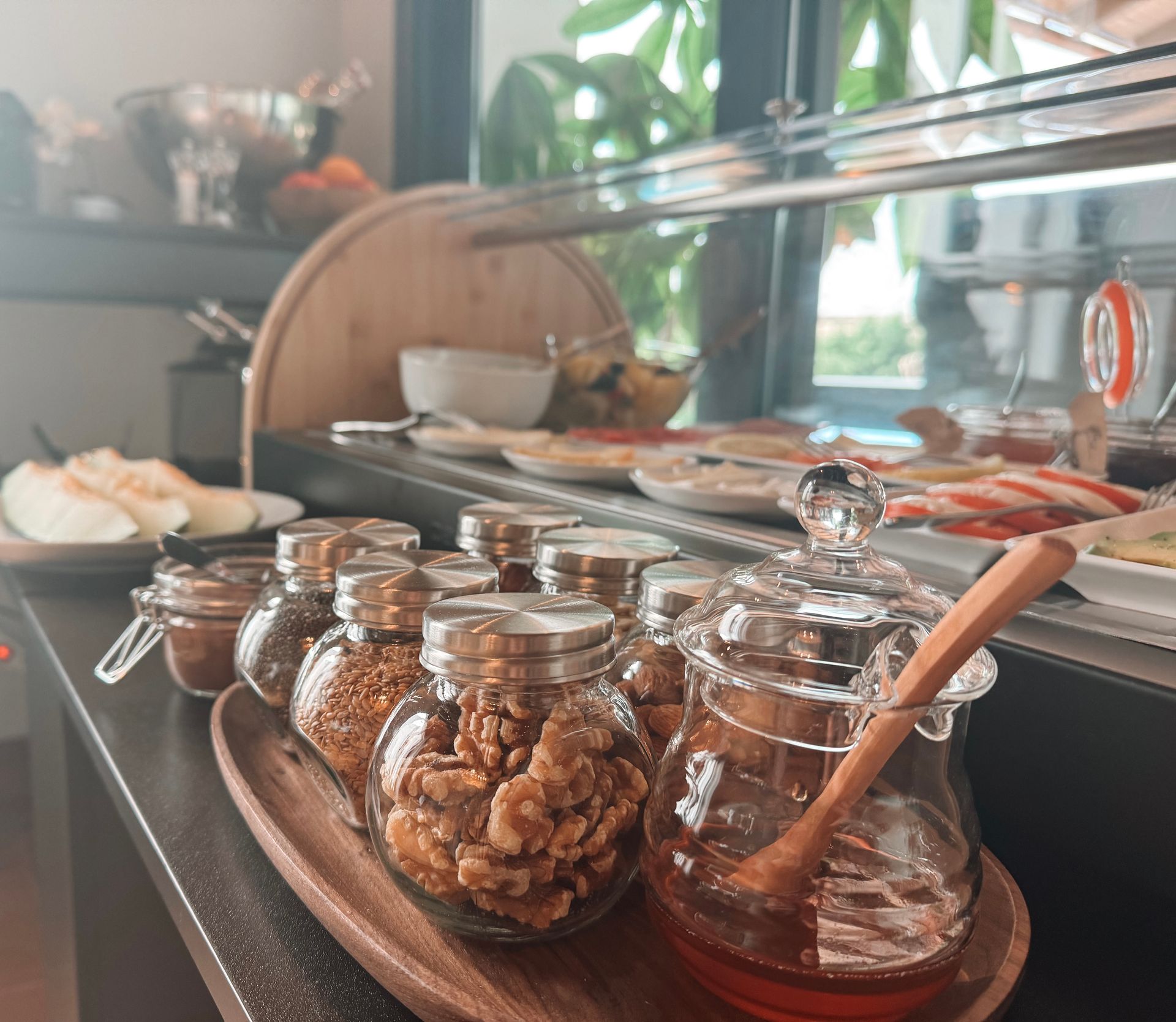 A buffet-style breakfast display with jars of honey, nuts, and spices, and fruit. A wooden serving tray sits in the foreground.