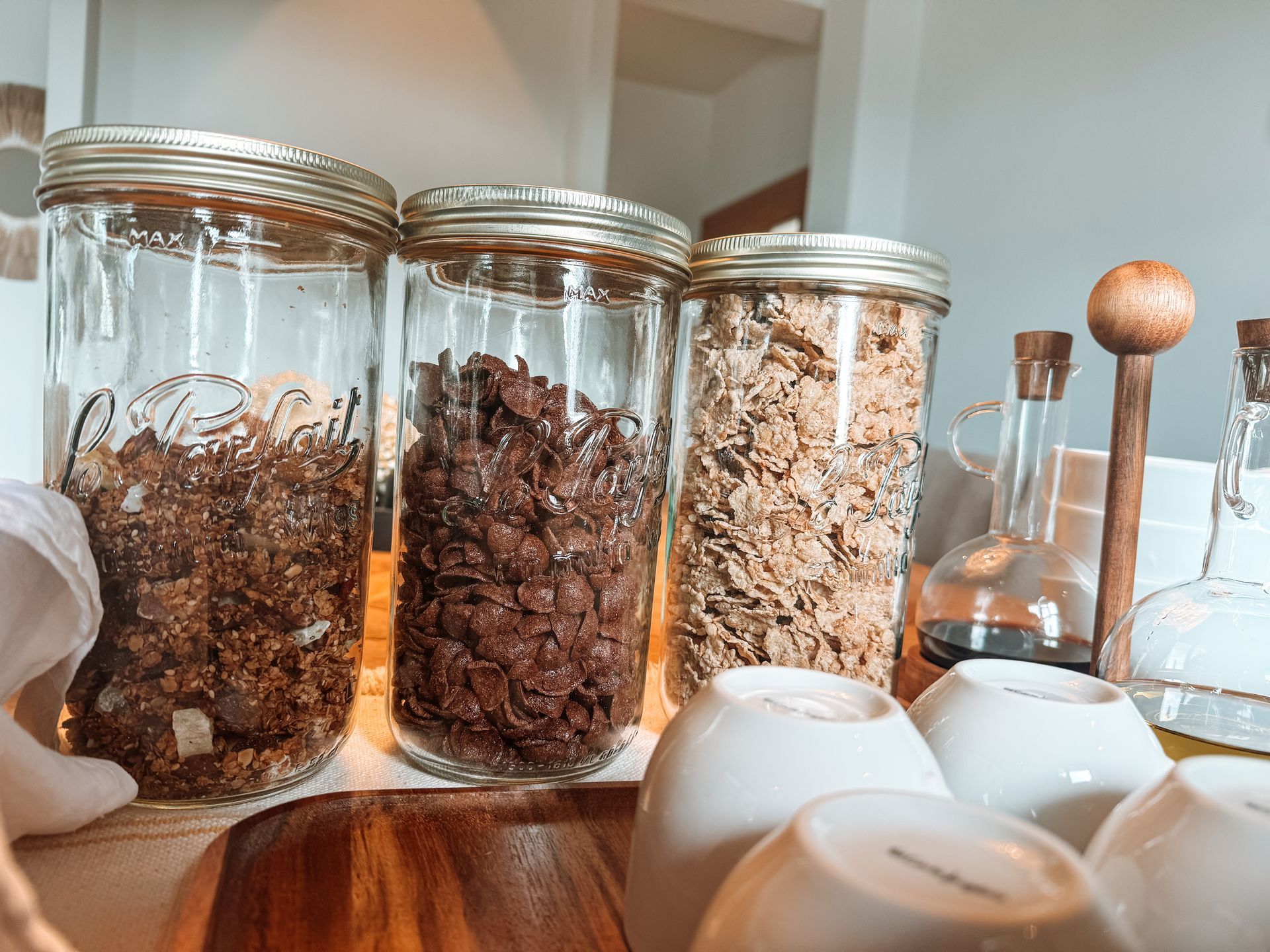 Three glass jars filled with cereal, next to stacked white bowls, on a wooden tray.