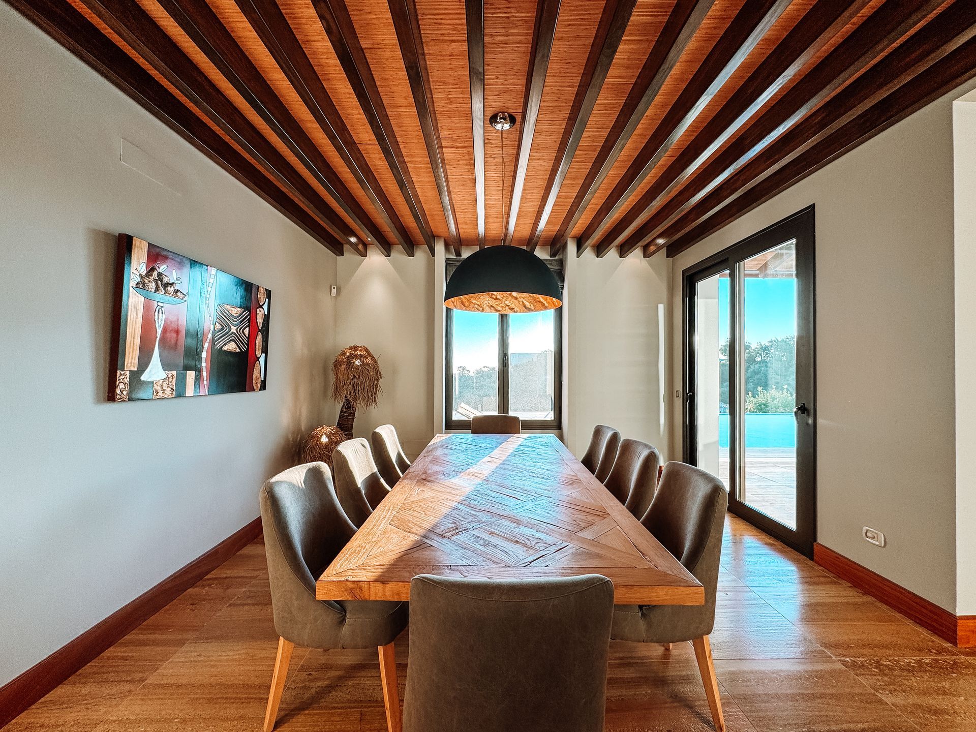 Dining room with a long wooden table, chairs, and a beamed ceiling. Sunlight streams in, illuminating the table.