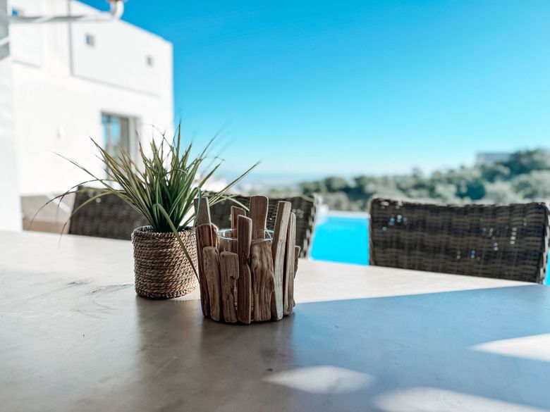 A sunny outdoor table with decorative items: a potted plant and a wooden candle holder. A blue swimming pool and sky are in the blurred background.