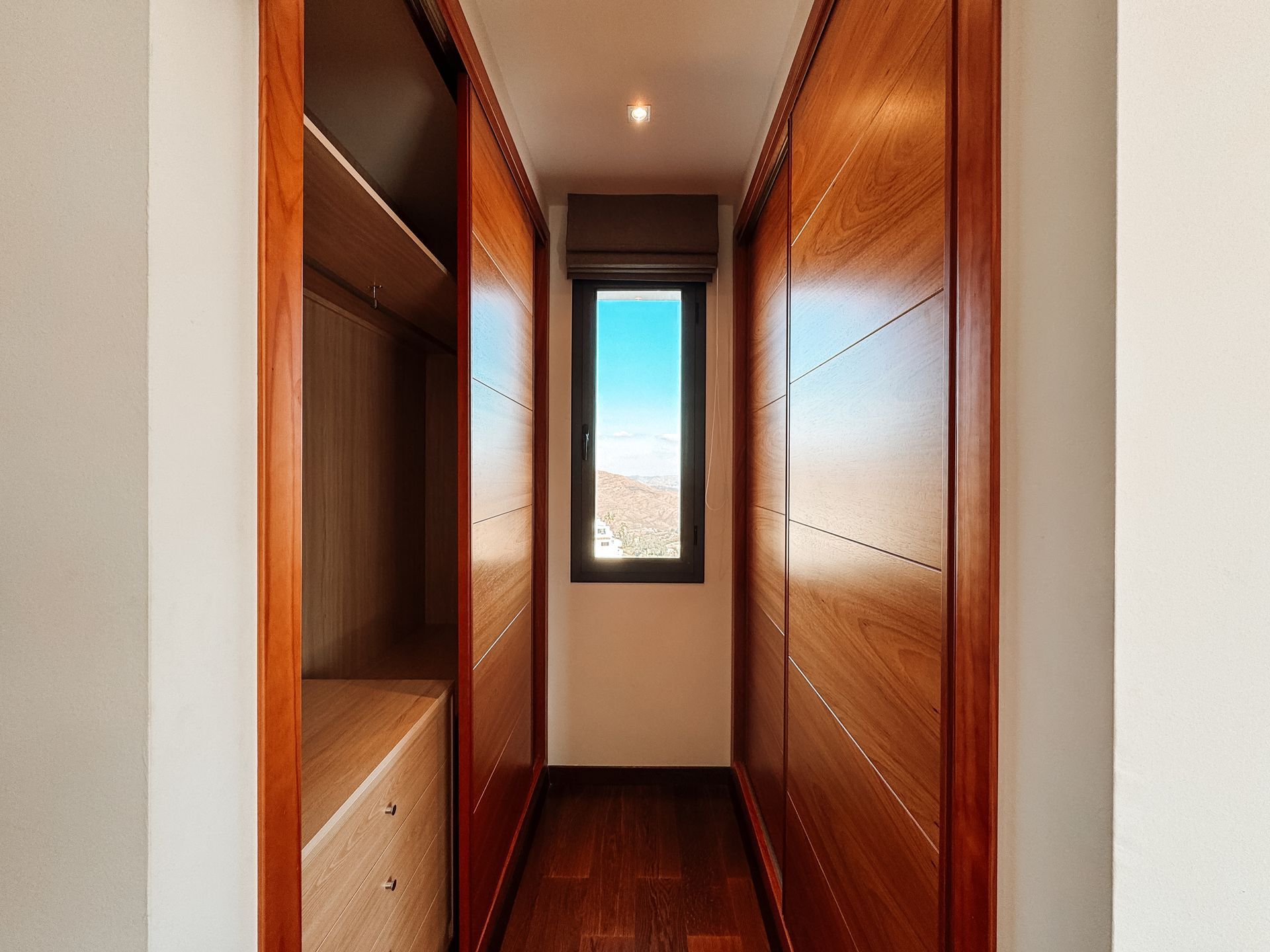 Narrow walk-in closet with dark wood doors and interior. A small window at the end offers a view of the outdoors.