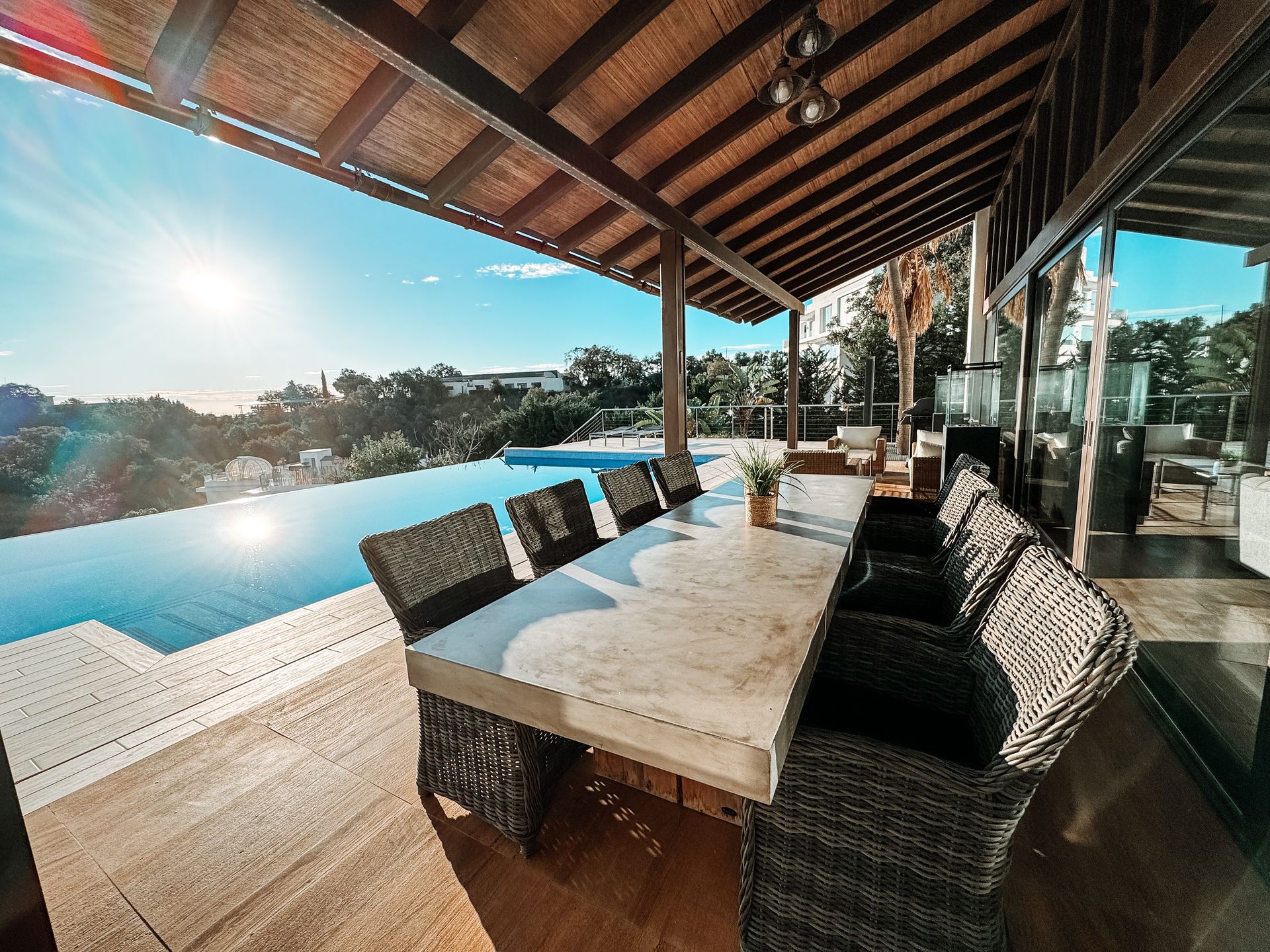 Outdoor dining area with a long table set for a meal, next to a swimming pool, under a wooden roof.