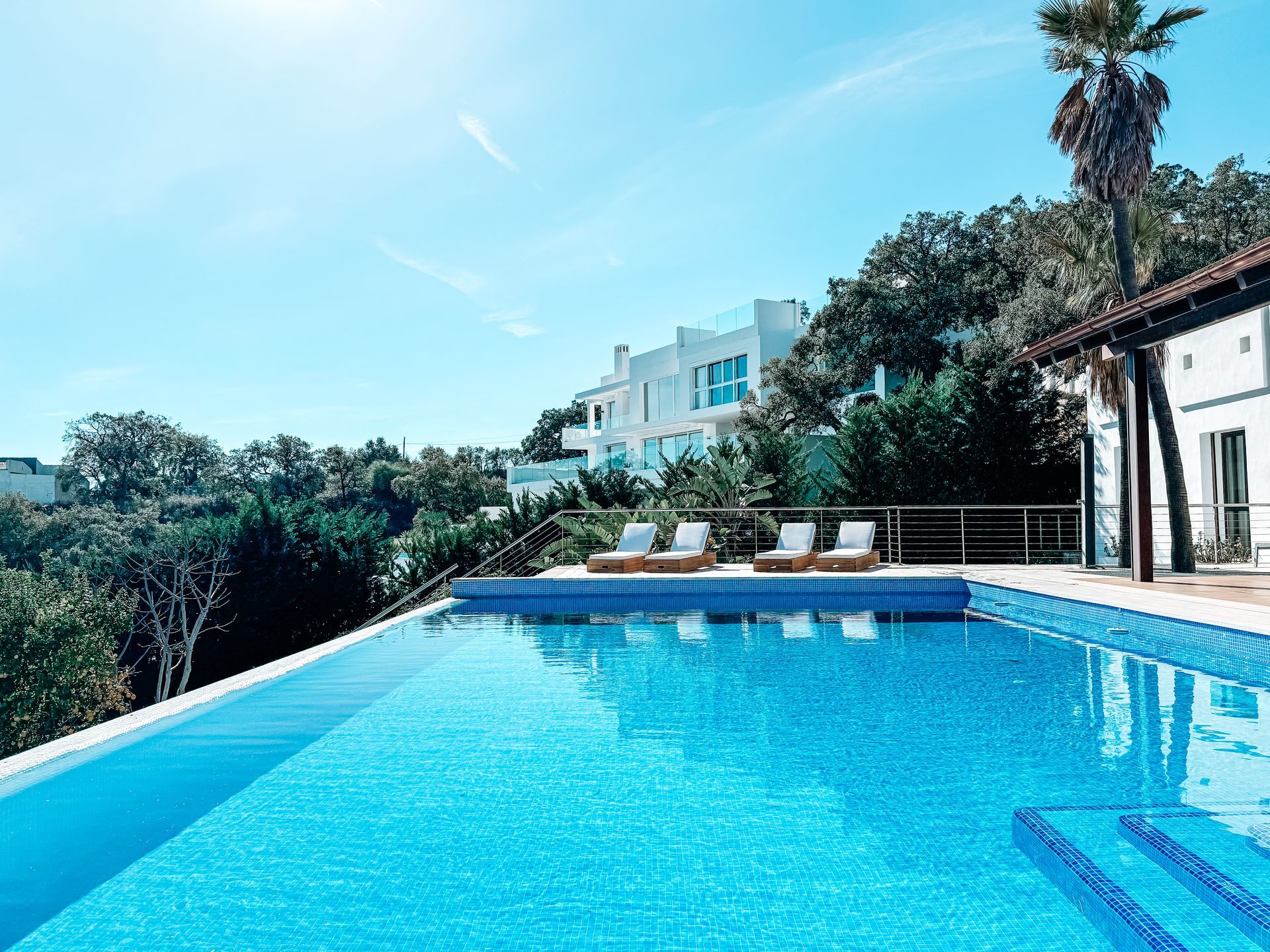 An infinity pool overlooking a sunny landscape, with lounge chairs, trees, and a white building in the background.