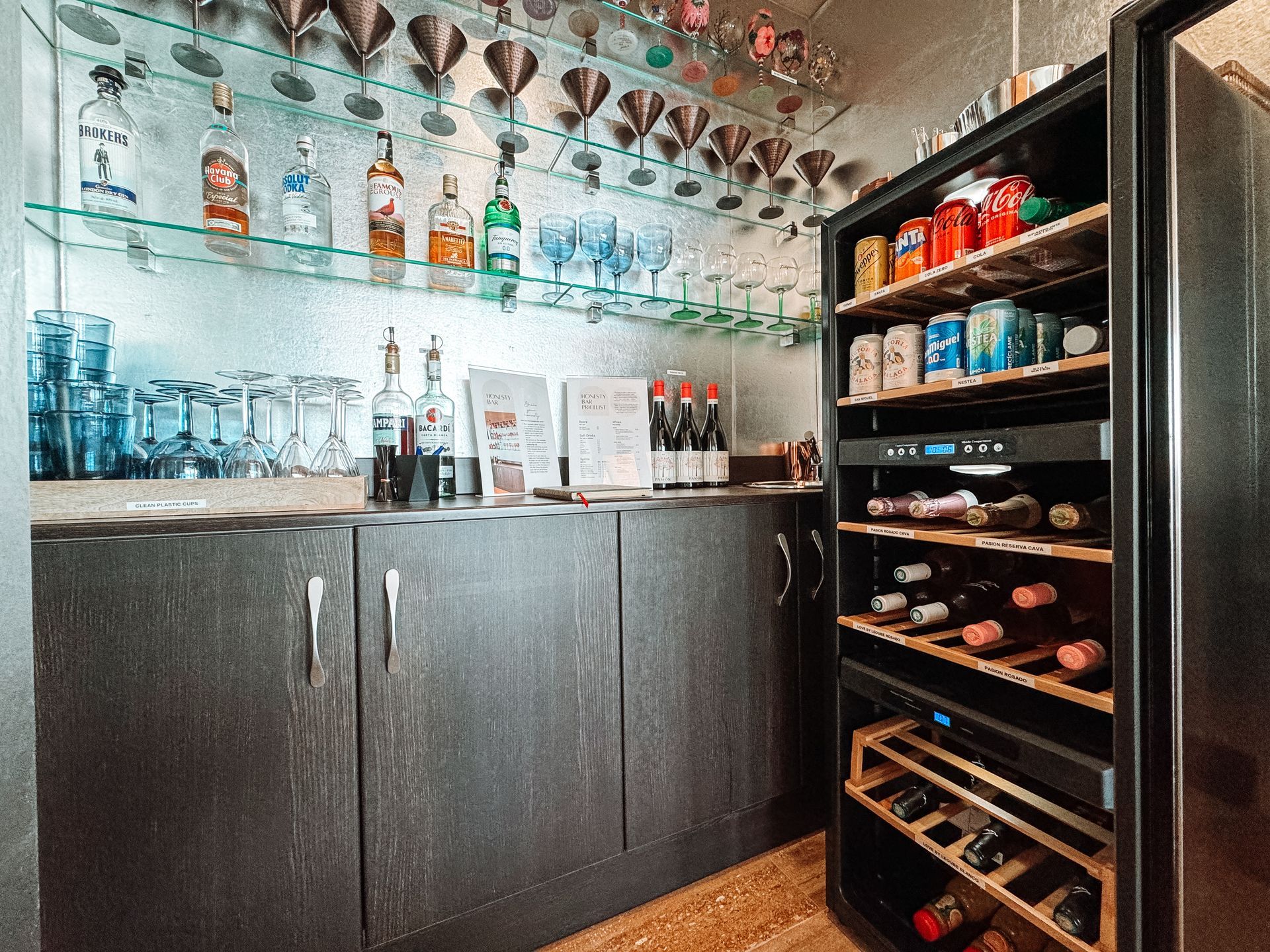 A bar with liquor bottles, glasses, and a wine cooler. The bar has dark cabinets, shelves, and a mirrored wall.