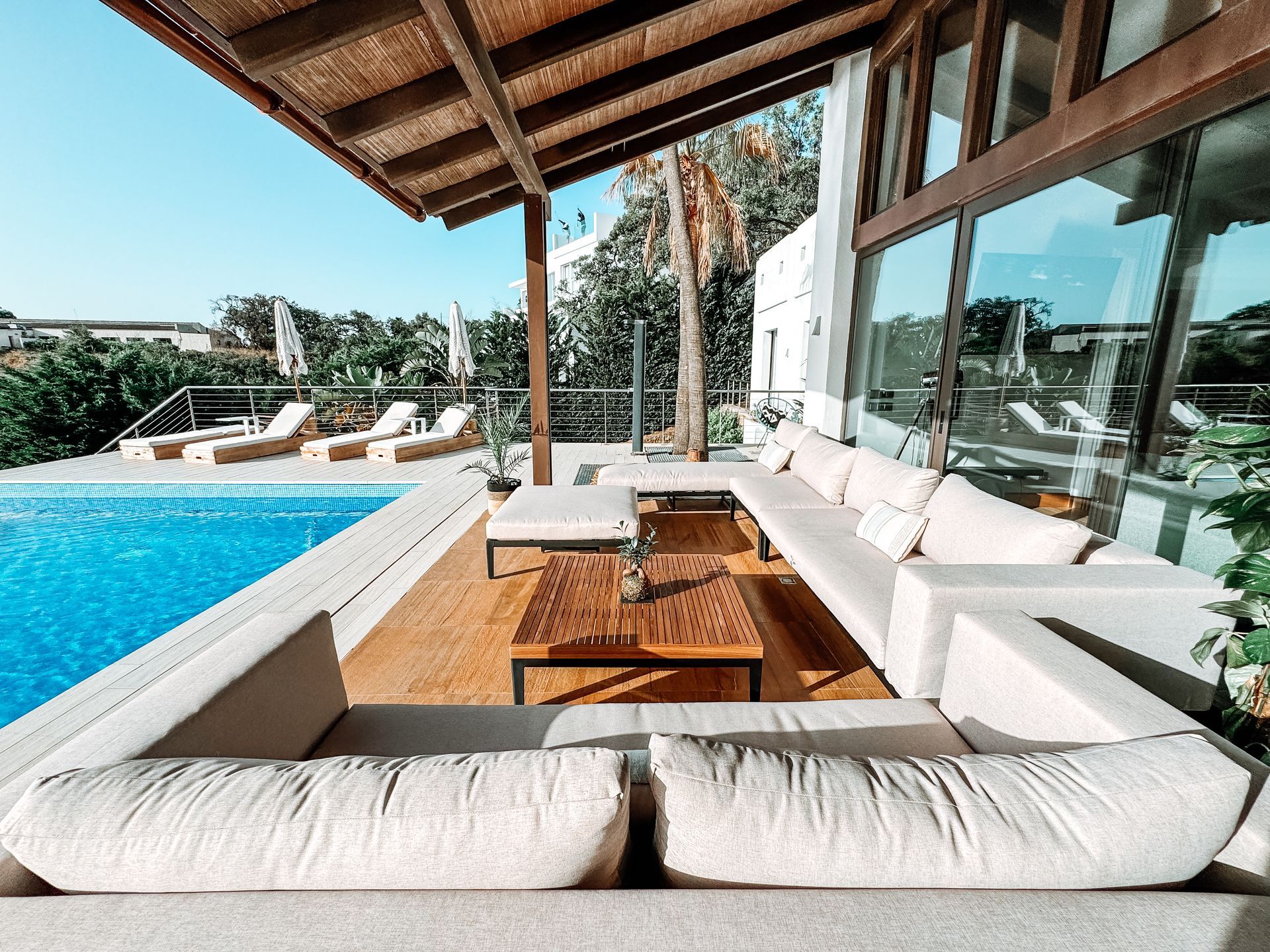 Outdoor living area with pool, beige sectional sofa, wooden tables, and glass windows. Sunny day.