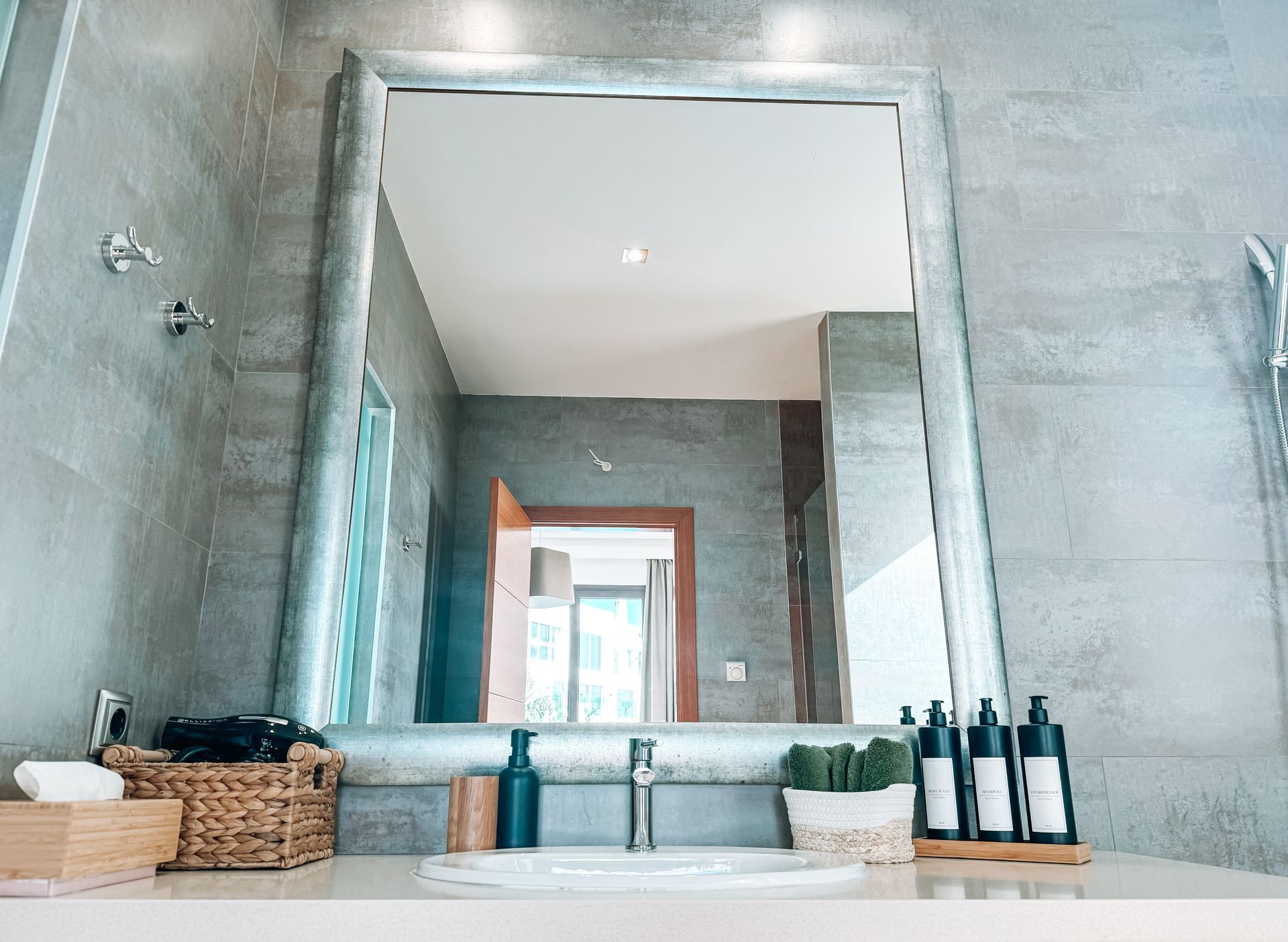 A large mirror above a bathroom sink reflects a doorway, gray walls, and toiletries. A wooden tray holds bottles.