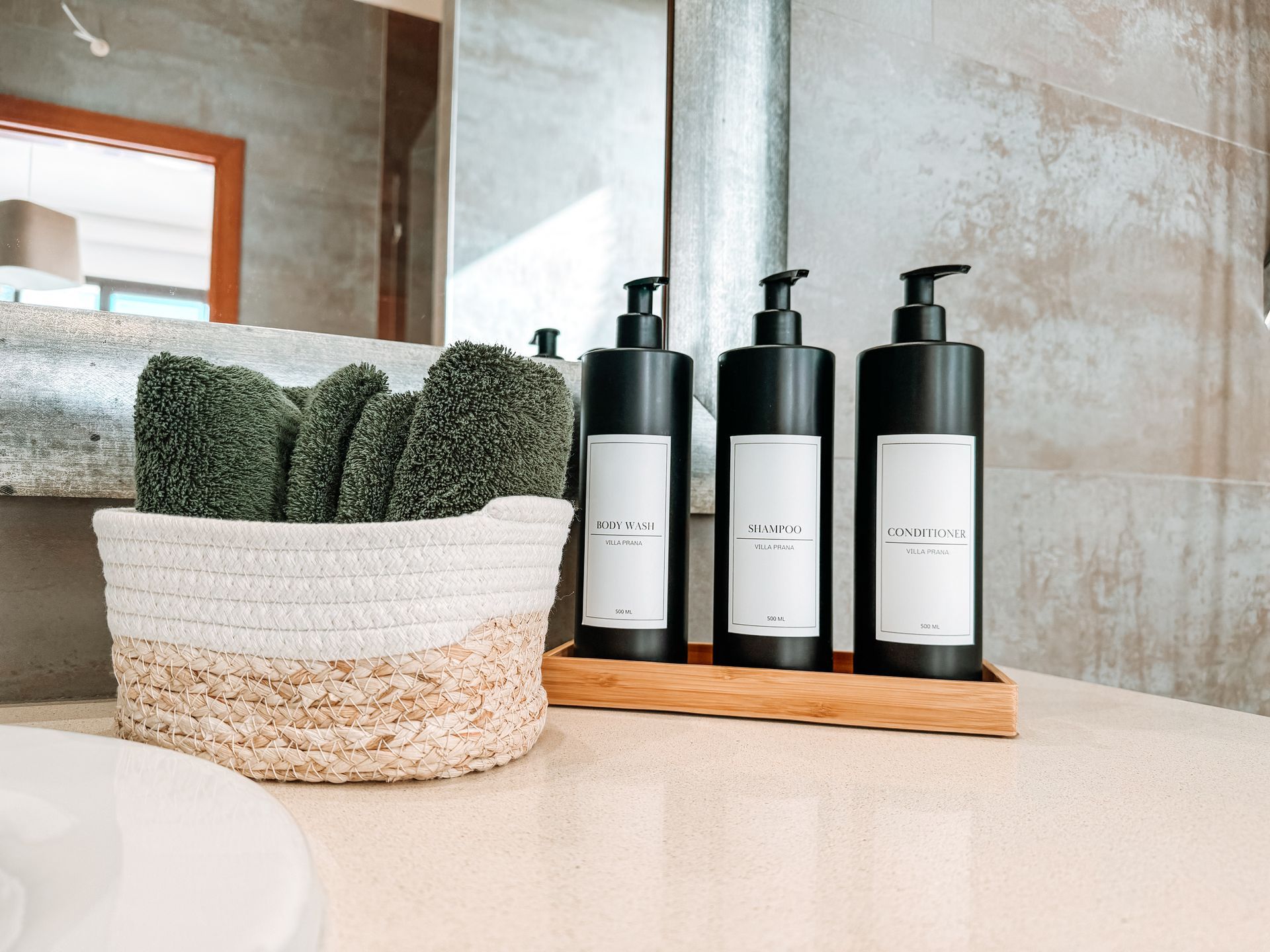 Bathroom counter with a basket of green washcloths, three black soap dispensers on a wood tray, and a mirror.