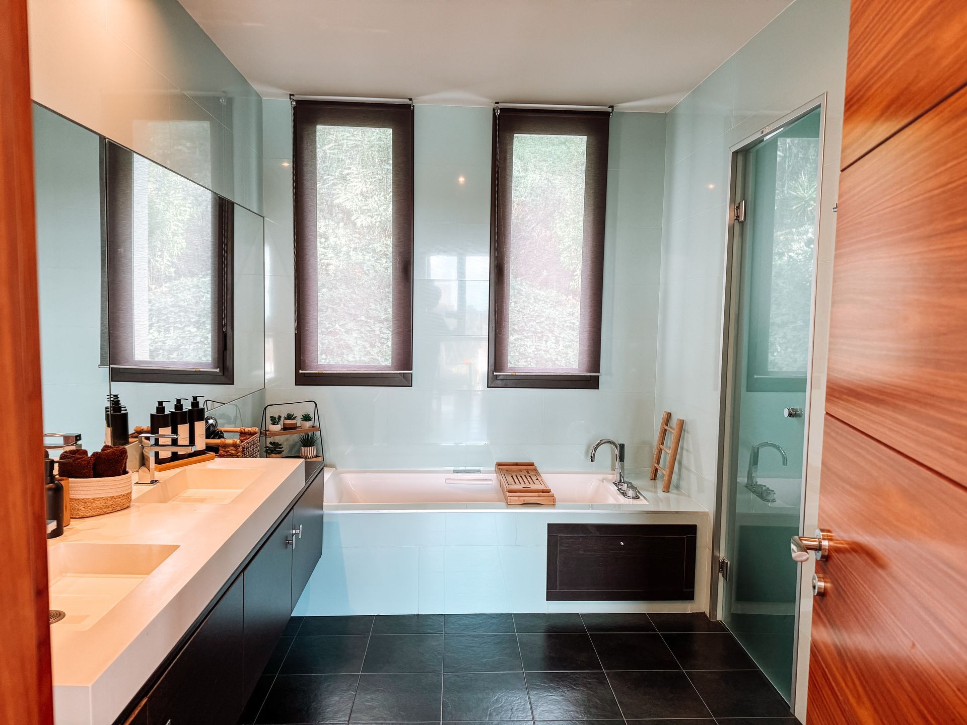 Modern bathroom with light blue tiled walls, dark floor, double vanity, and a bathtub with wooden accents.