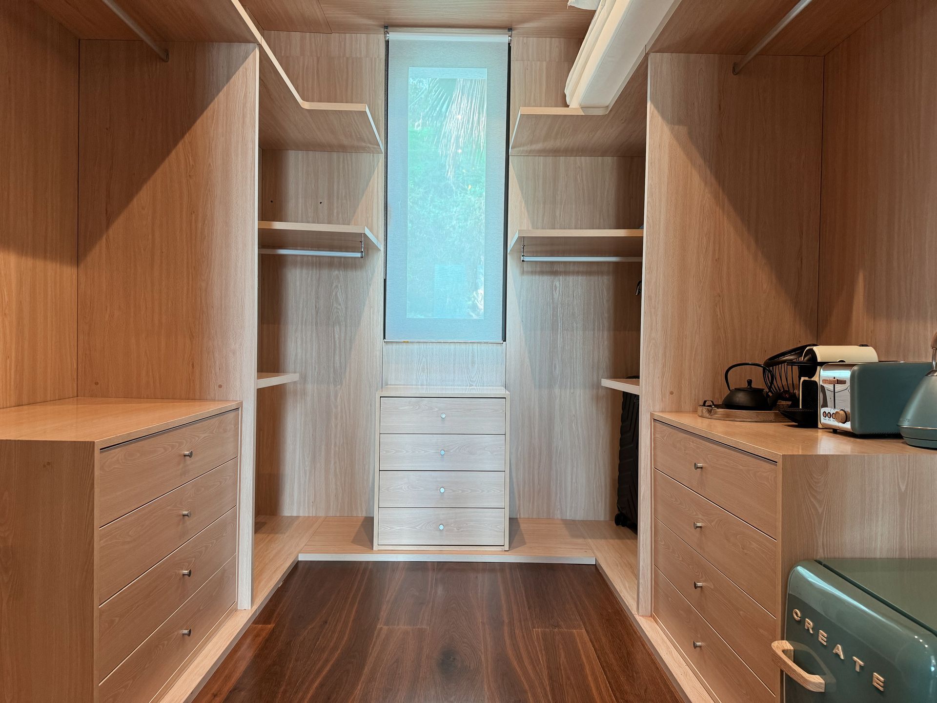 A light-wood walk-in closet with built-in drawers, shelves, and hanging rods. A narrow window is centered on the back wall.