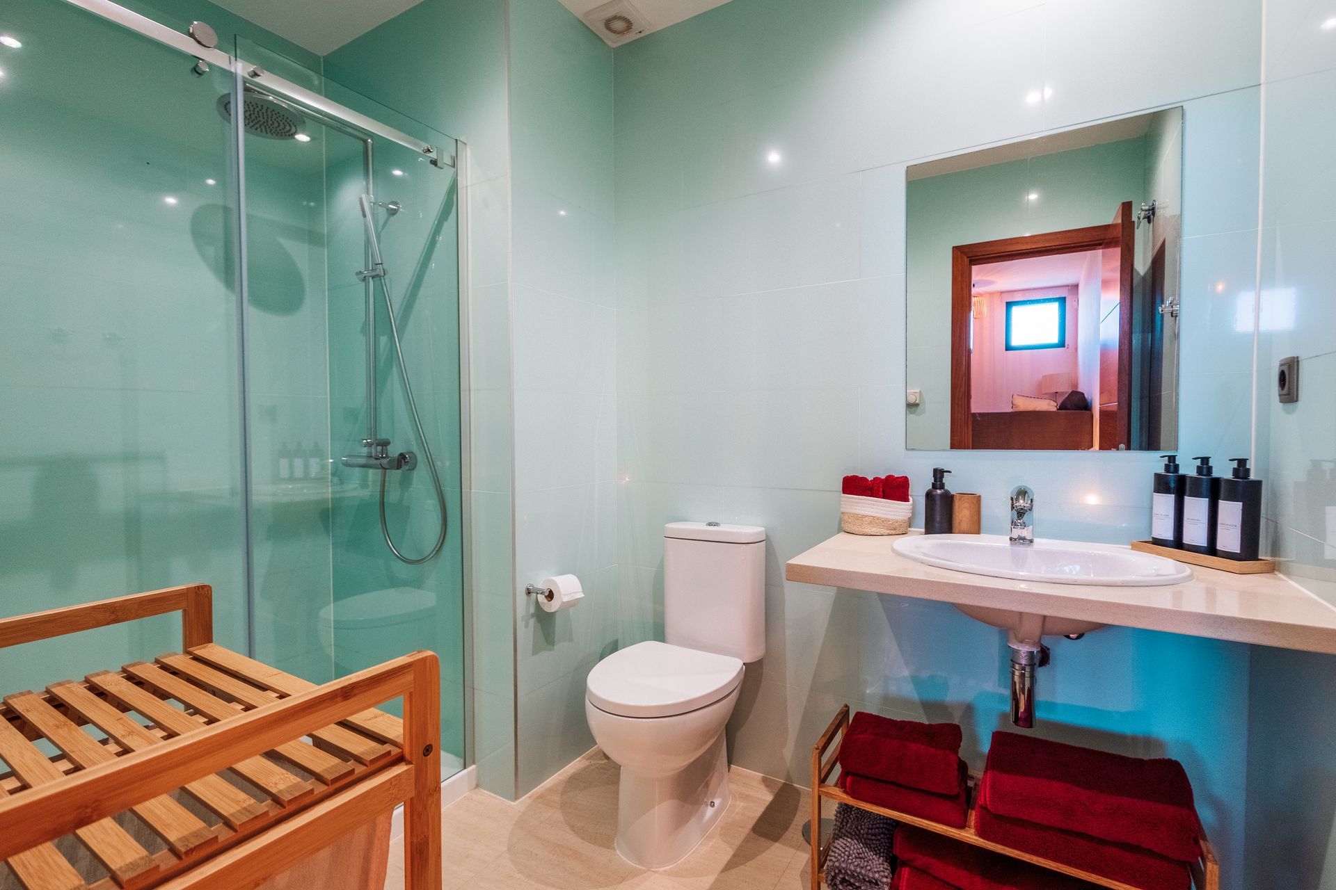 A light blue bathroom features a glass shower, toilet, sink, and wooden bench. The mirror reflects a glimpse of another room.