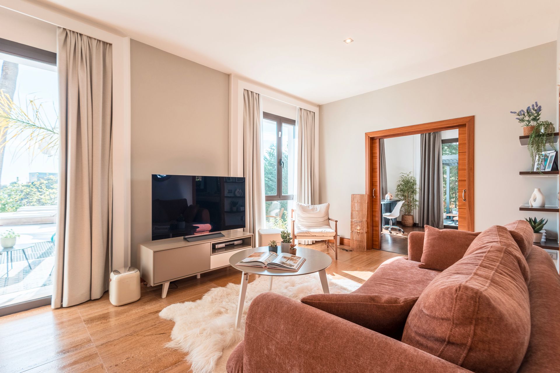 Cozy living room with hardwood floors, a pink sofa, large windows with light-colored curtains, and a mounted TV.