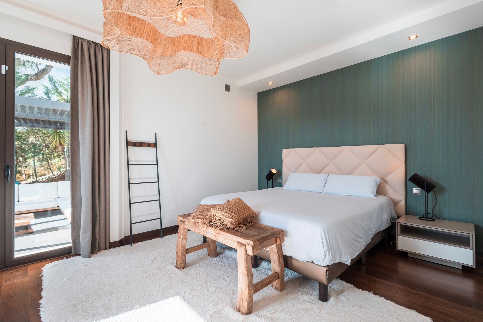 Bedroom with a bed, a green accent wall, and a large rug. There is a wooden bench at the foot of the bed and a light fixture above.