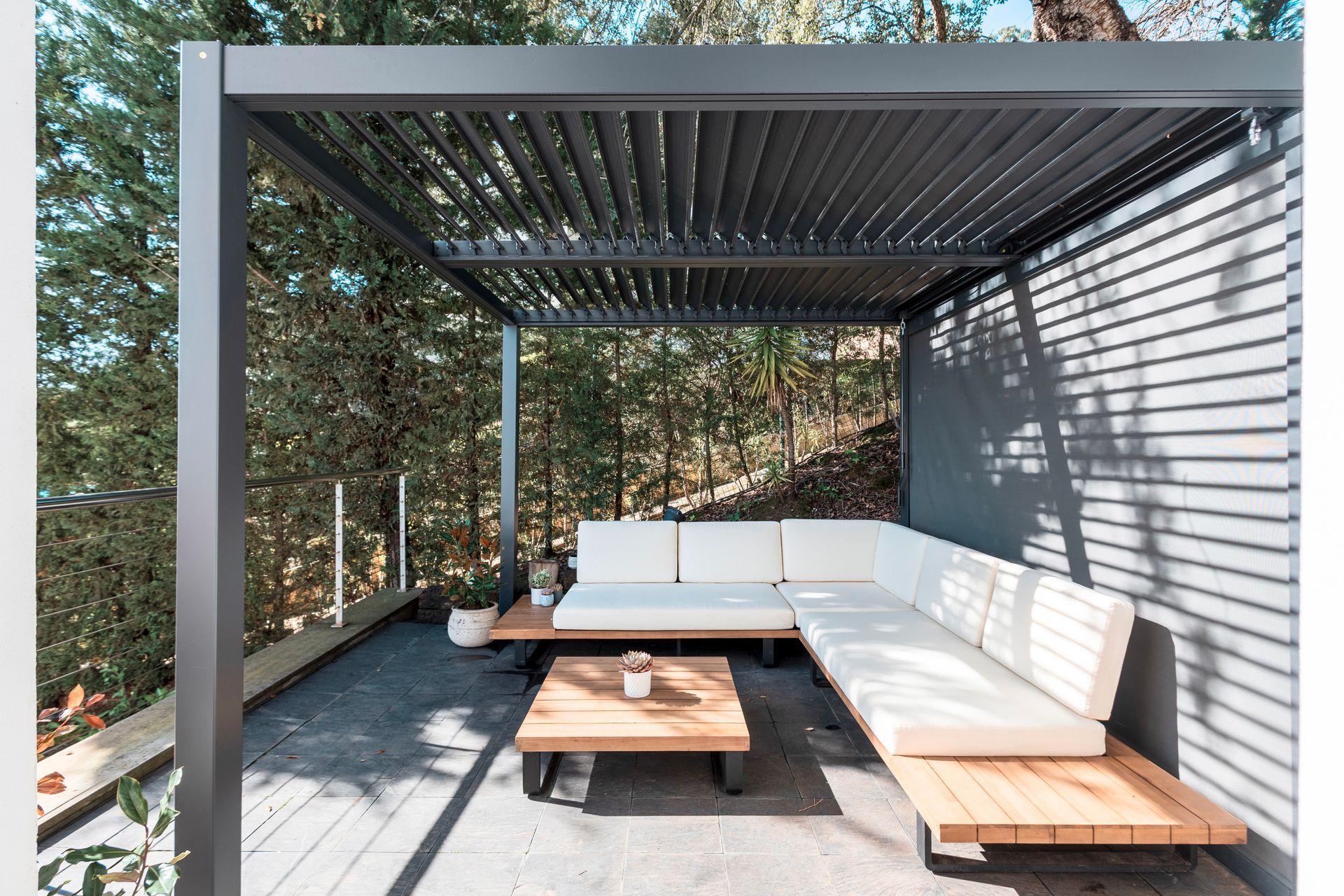 Outdoor seating area with a modern pergola, a wooden sectional sofa with white cushions, and a wooden coffee table. The setting is next to a forested area.