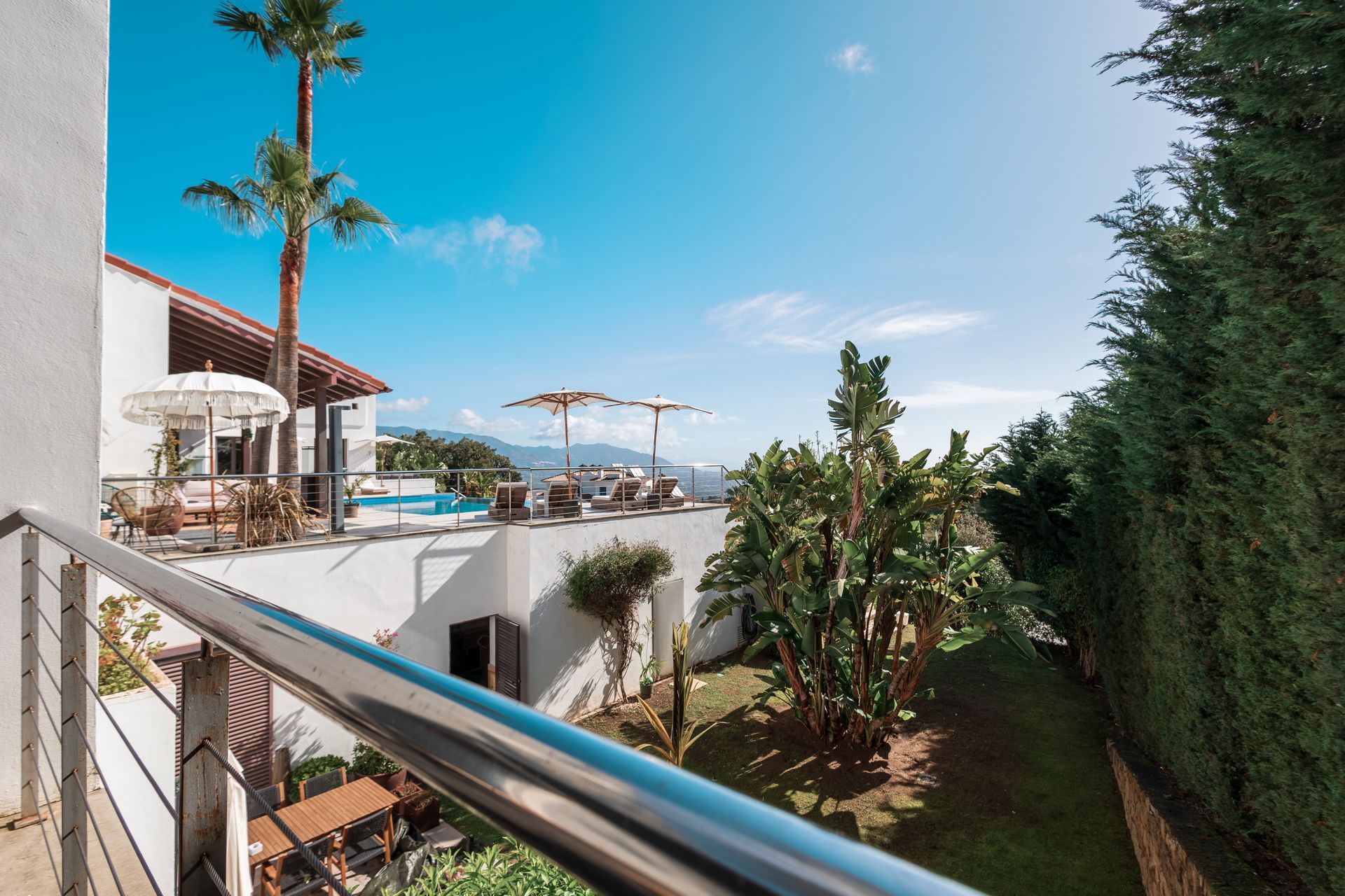 View from a balcony overlooking a white villa with a pool, palm trees, and blue sky.