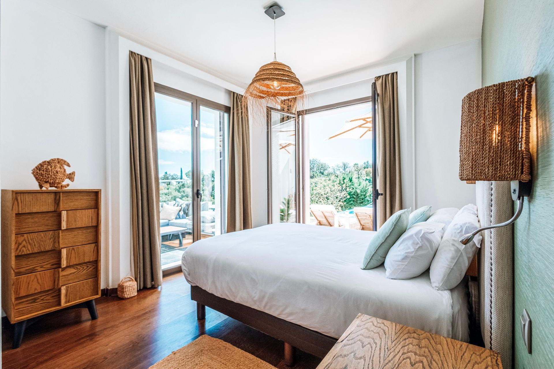 Bedroom with a bed, wooden furniture, and a view through open glass doors onto a terrace with greenery.