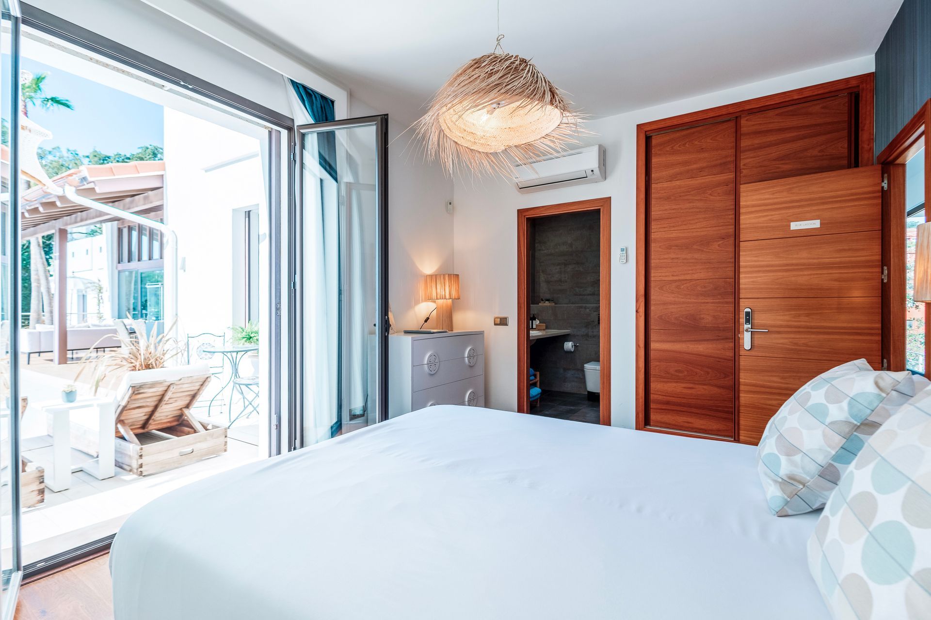 Bedroom with a white bed, open doors to a patio, wooden wardrobe, and a small bathroom in view.
